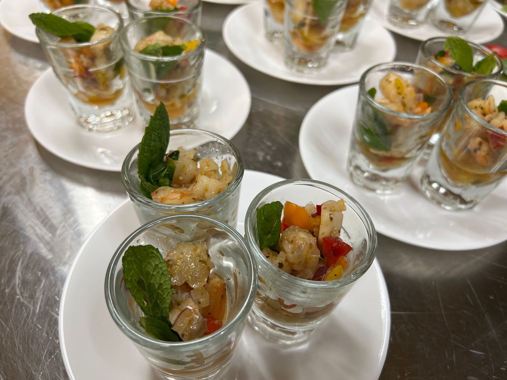 Small shot glasses filled with shrimp ceviche, garnished with mint leaves and set on white plates on a metal surface.