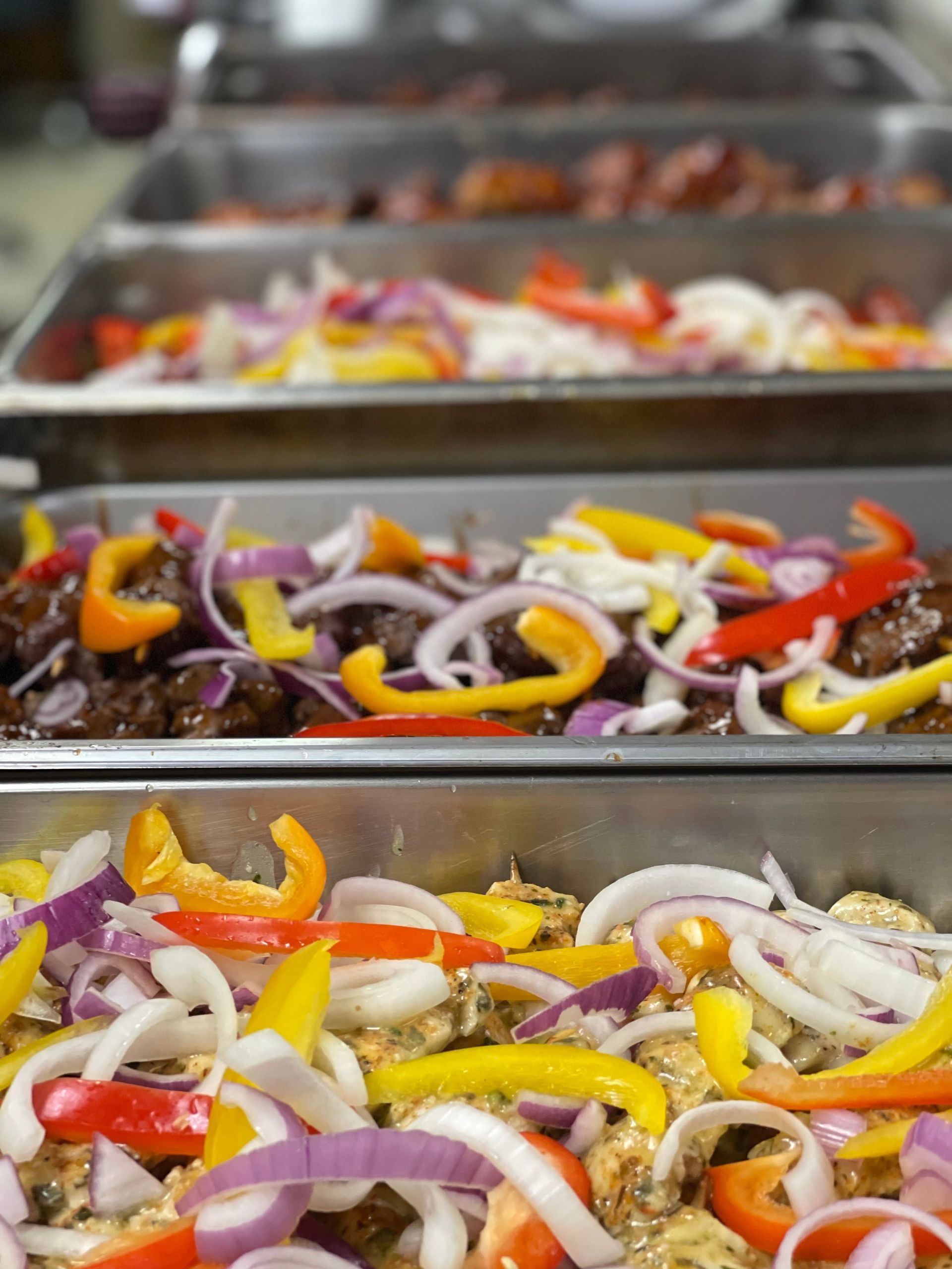 Several metal catering trays filled with colorful sliced bell peppers, red onions, and meat prepared for a buffet.