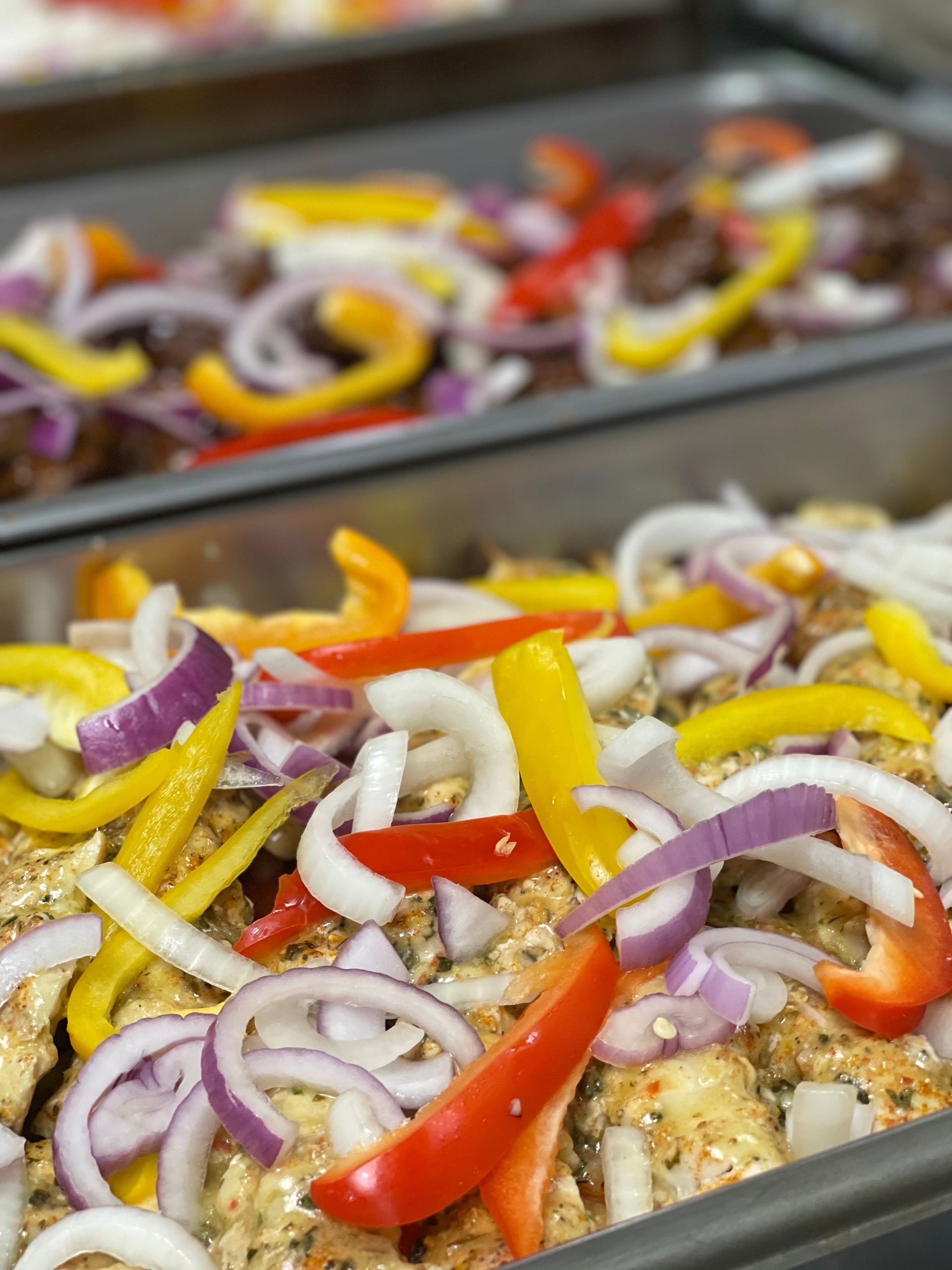 Metal serving trays containing marinated chicken topped with sliced red onion, yellow bell pepper, and red bell pepper.