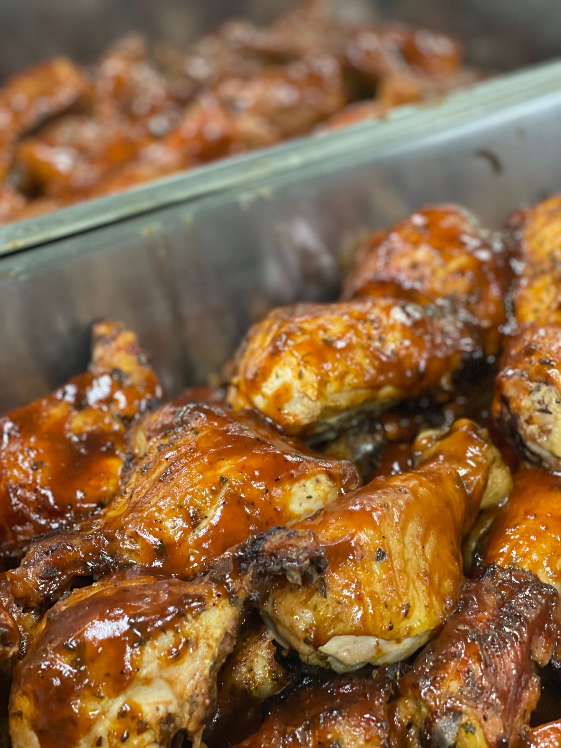 Close-up of two metal catering trays filled with golden-brown chicken pieces coated in a glossy, dark barbecue sauce.