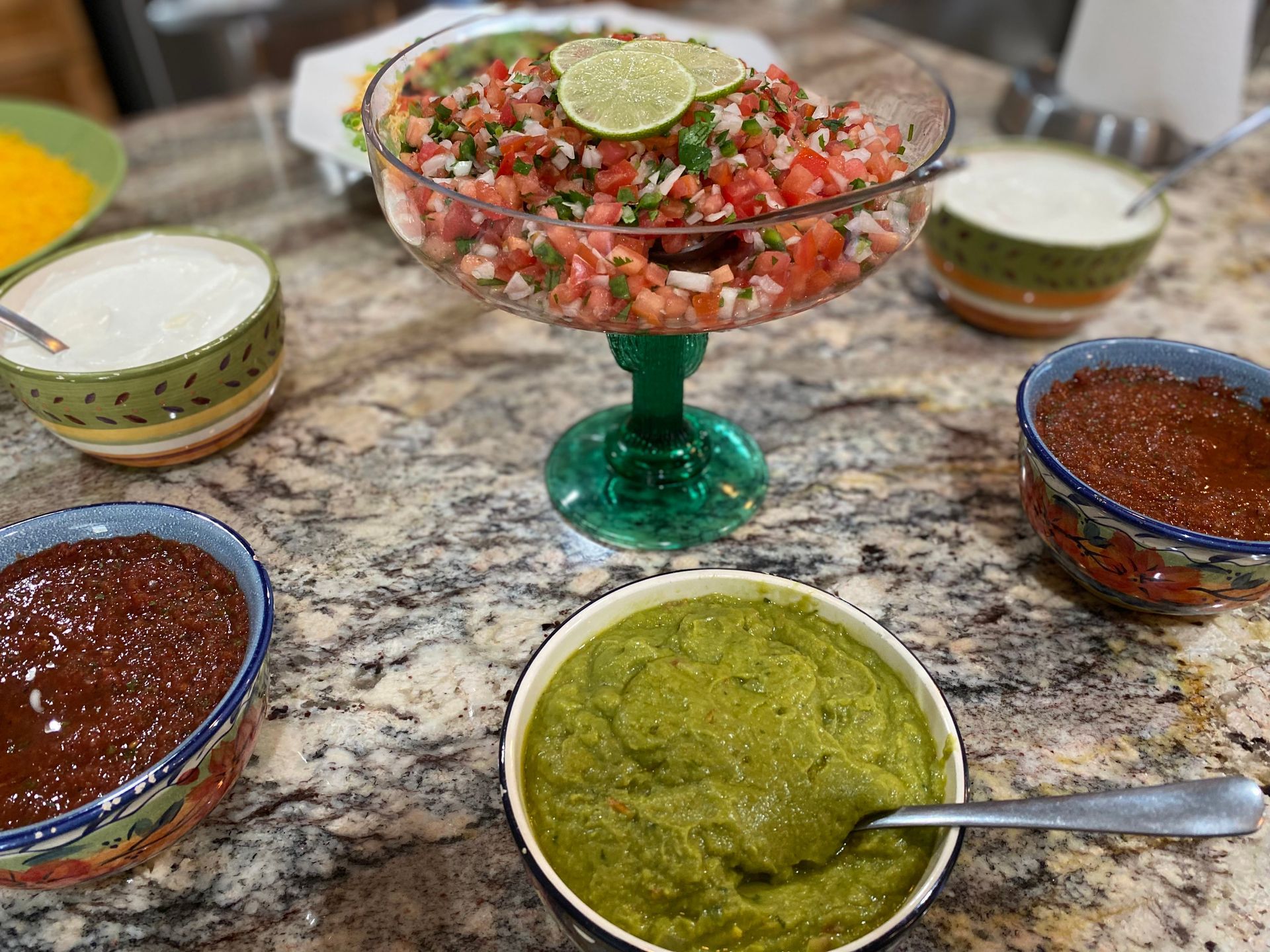 A festive taco bar spread featuring pico de gallo, salsa, and white sauce in colorful bowls on a granite countertop.