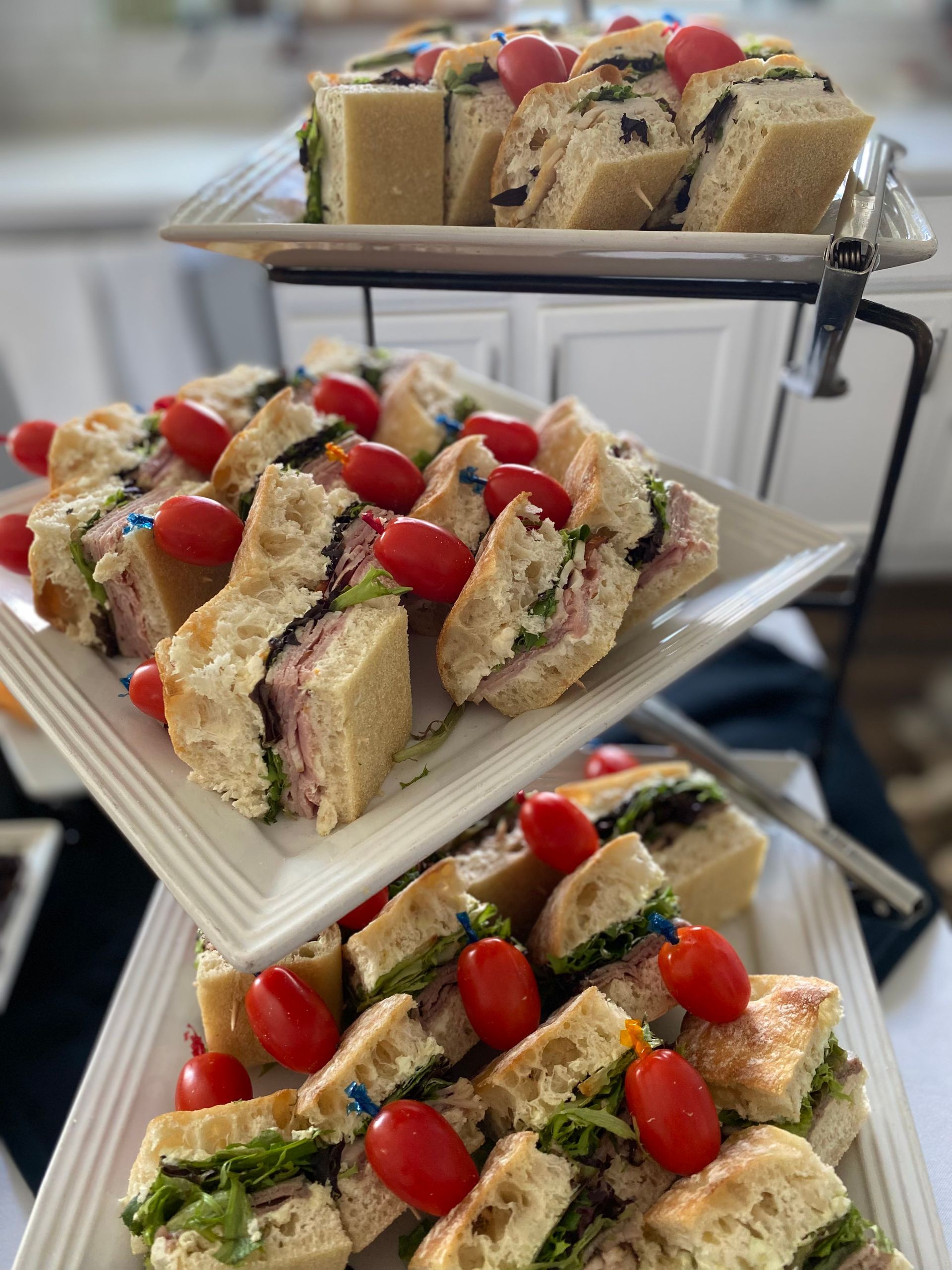 A three-tiered serving stand displays rows of focaccia sandwiches, each topped with a fresh cherry tomato and a toothpick.