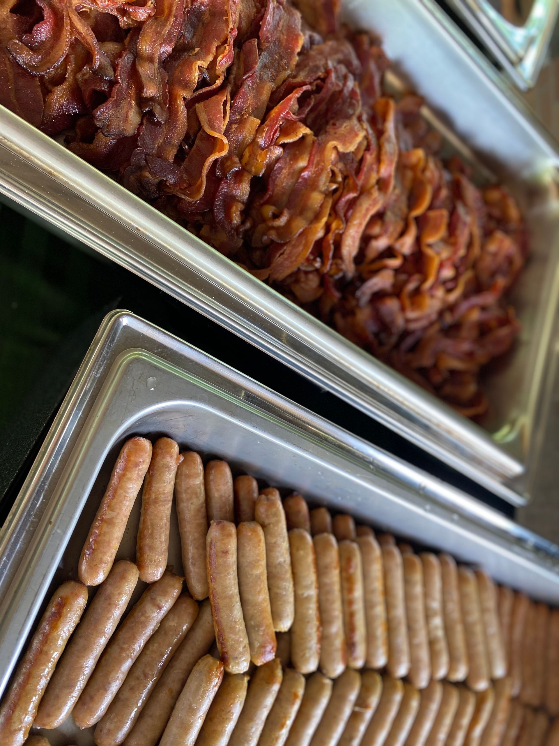 Two metal catering trays filled with cooked bacon strips and breakfast sausage links.
