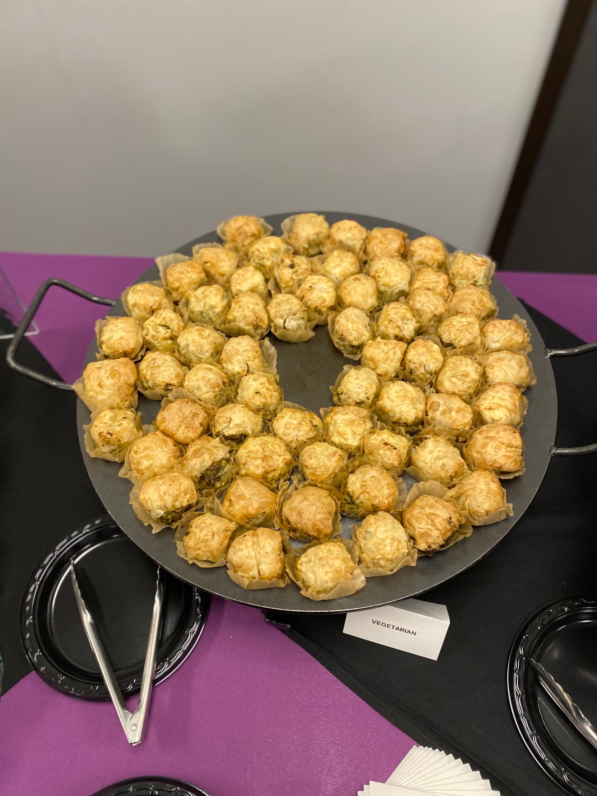 A round tray filled with bite-sized, golden-brown baked appetizers on a table with black plates and purple placemats.