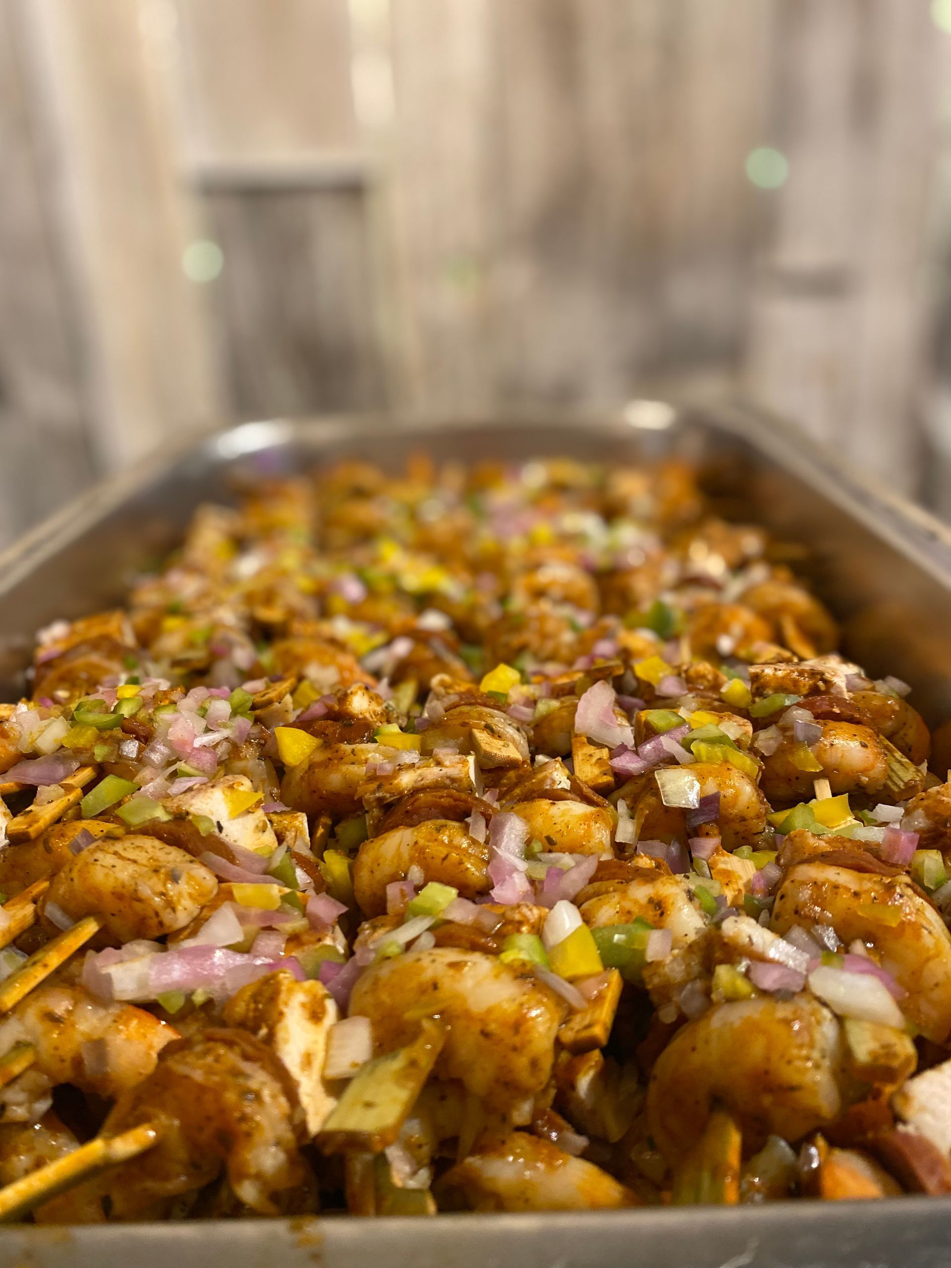 A metal catering tray filled with grilled shrimp skewers topped with diced red onions and yellow and green peppers.