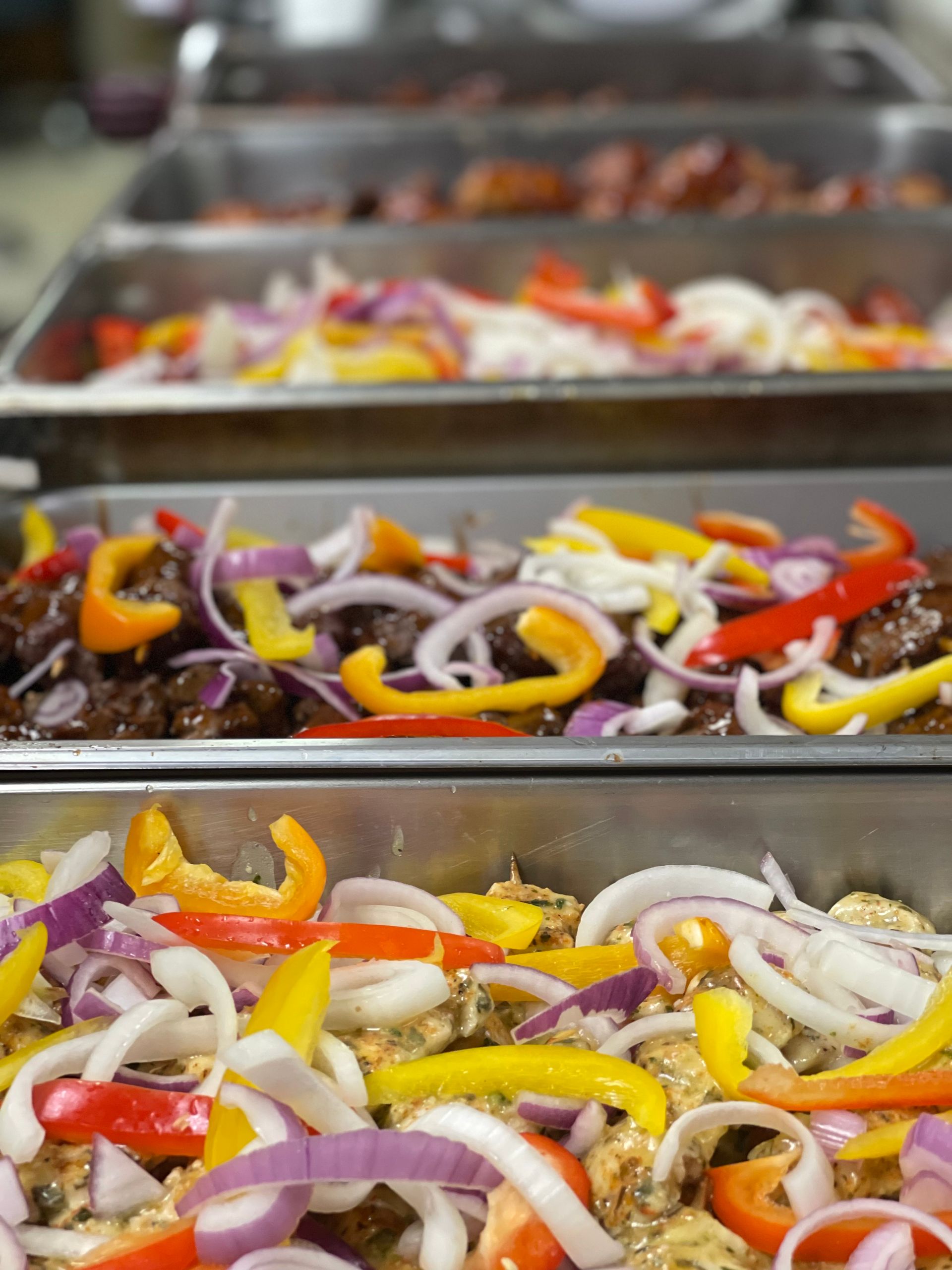 Rows of metal buffet pans filled with cooked meat topped with colorful sliced bell peppers and purple onions.