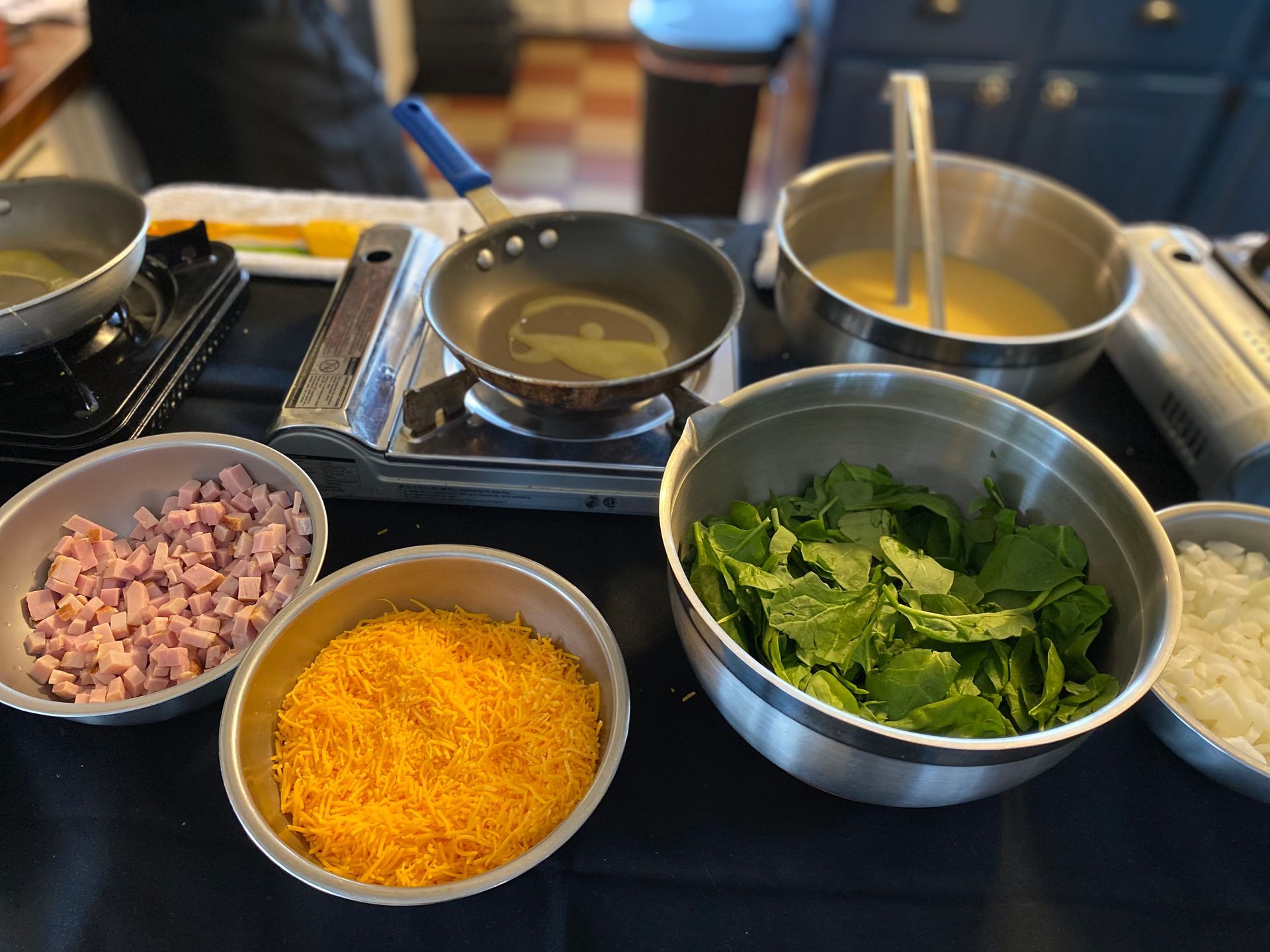 A station for making omelets with bowls of diced ham, shredded cheese, spinach, onions, egg mixture, and a hot skillet.