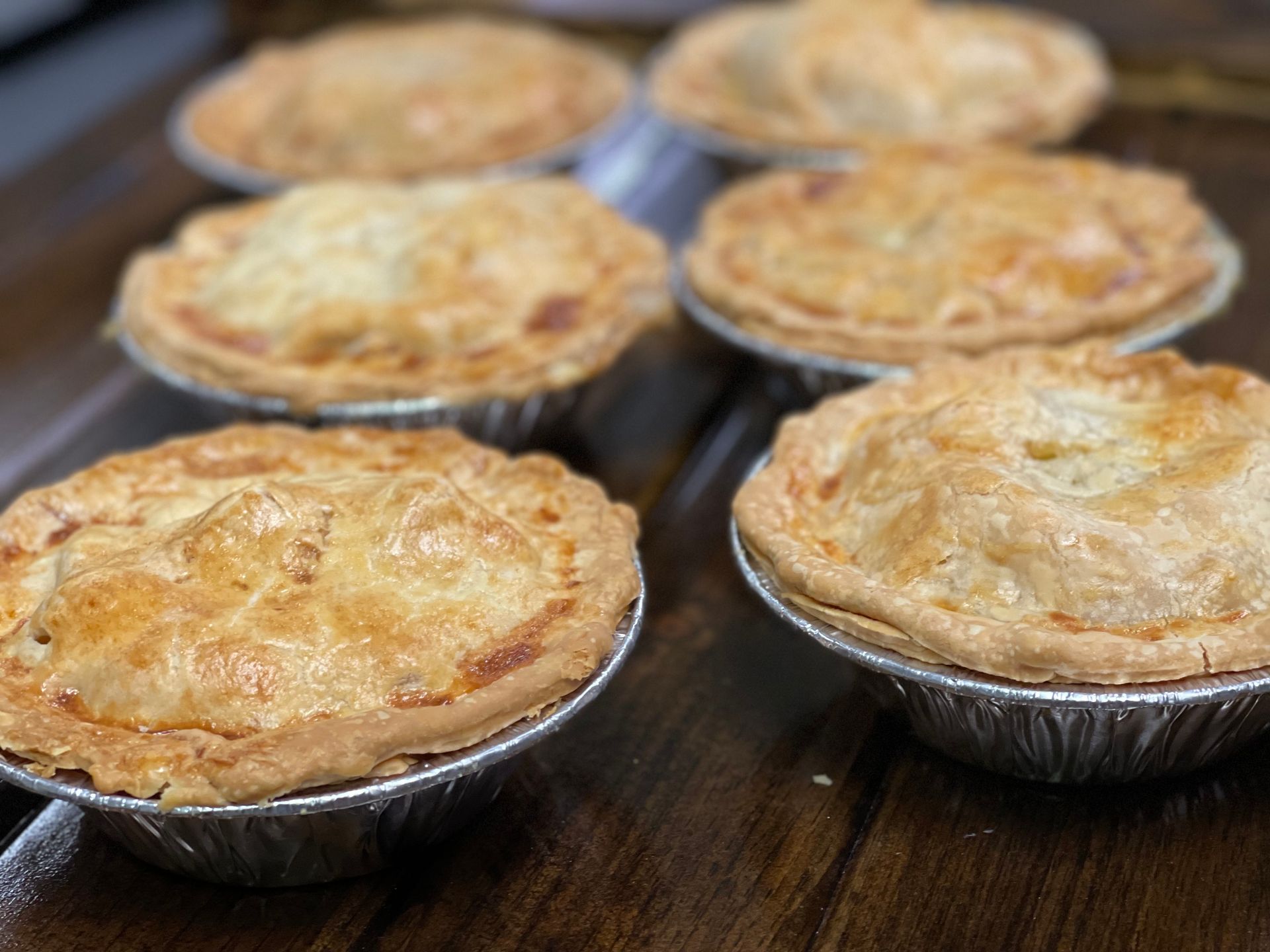 A group of six golden-brown savory pies in individual foil tins sitting on a wooden surface.
