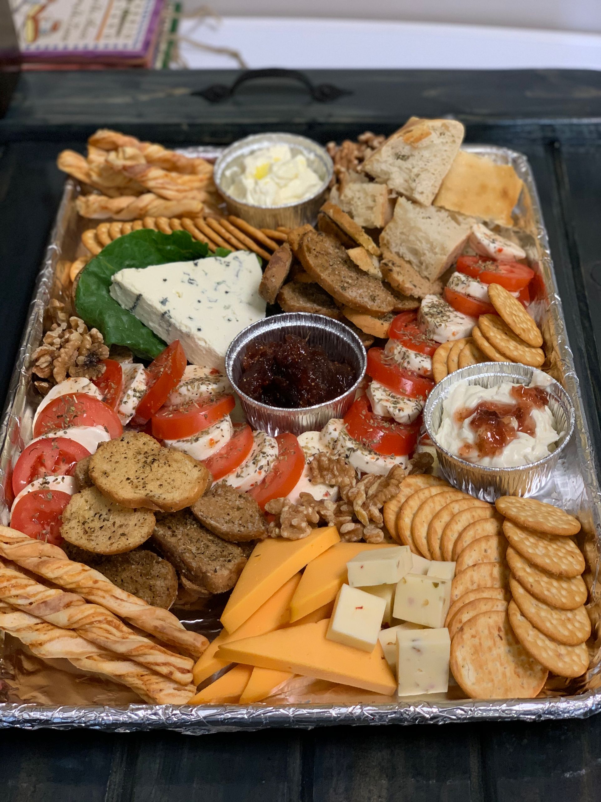 A charcuterie board featuring crackers, bread, cheeses, tomato slices, nuts, and bowls of spreads on a foil-lined tray.