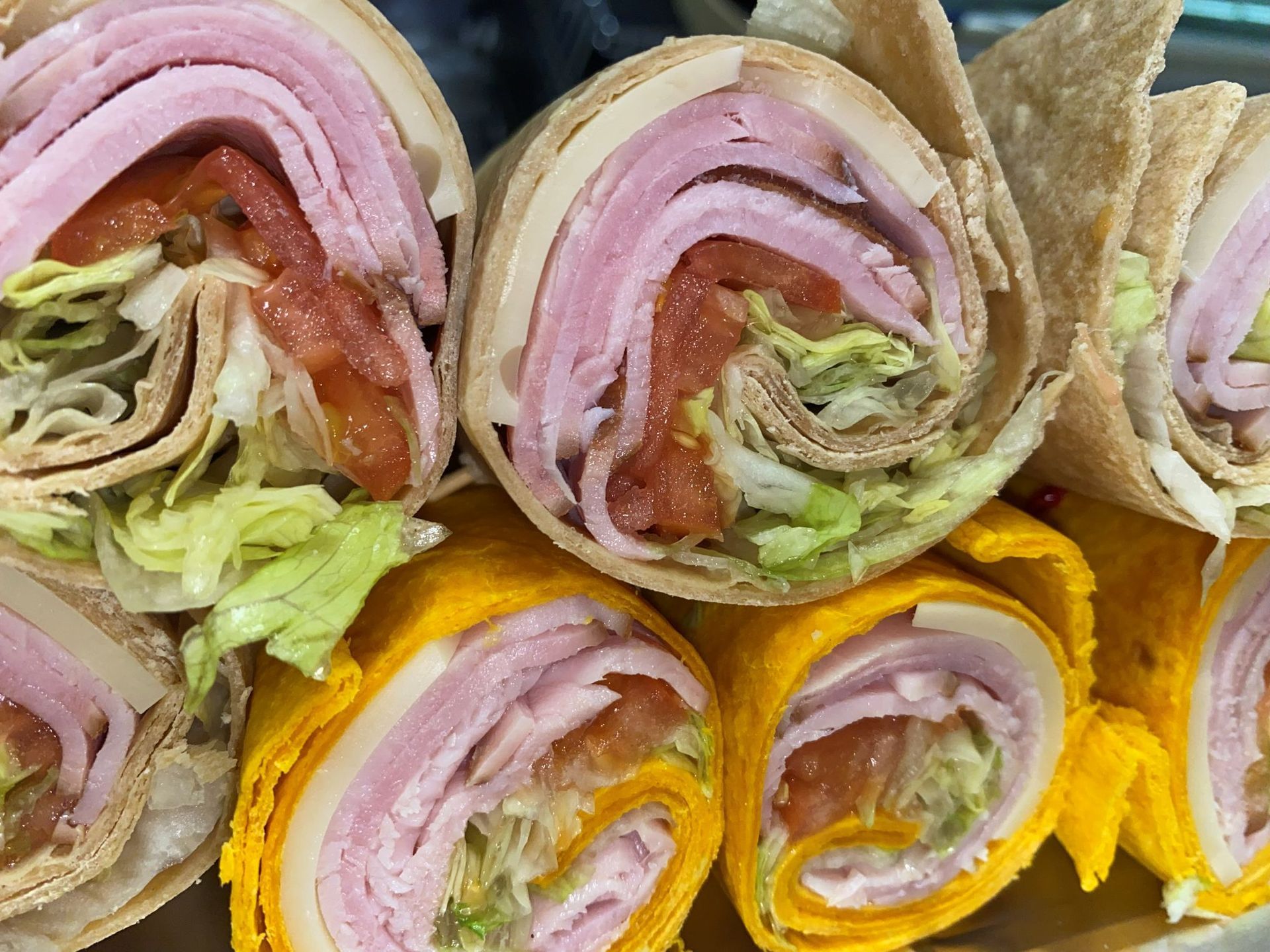 Wraps filled with ham, tomato, lettuce and cheese; some with yellow and some with brown tortillas.