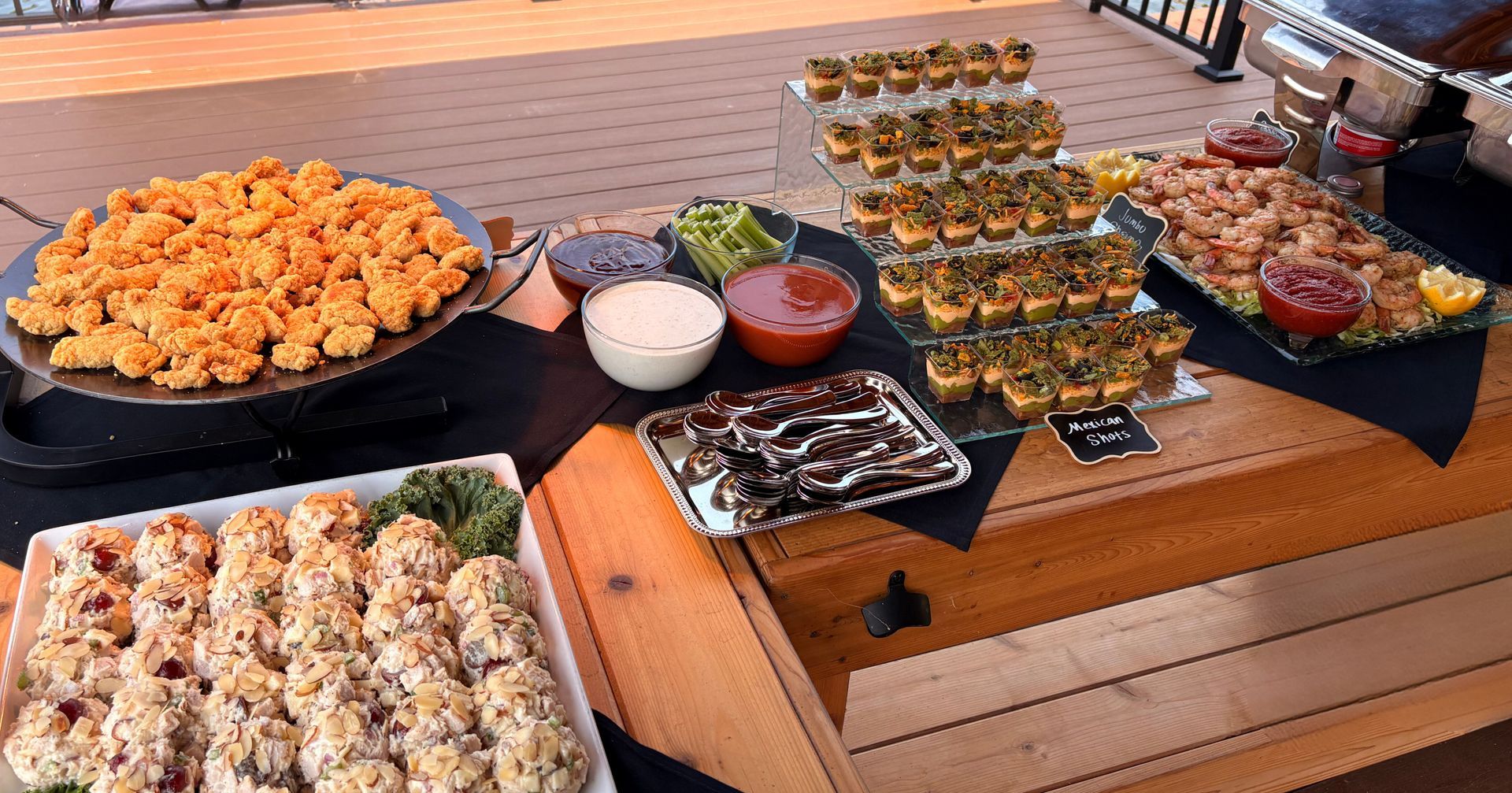 A buffet spread on a wooden table featuring fried food, shrimp, small appetizers on a tiered stand, and sauces.