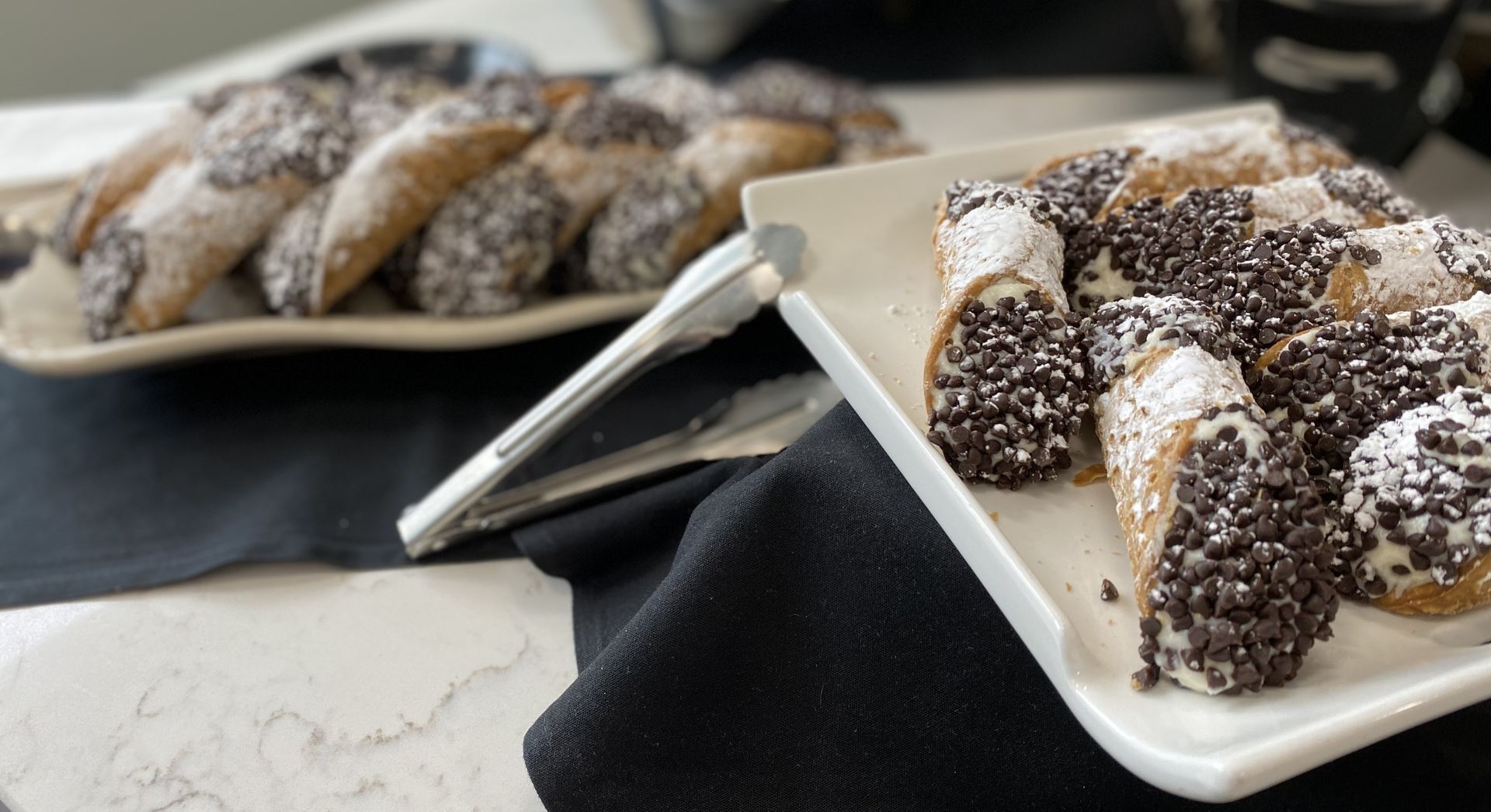 Cannoli with chocolate chips and powdered sugar served on white rectangular plates with serving tongs.