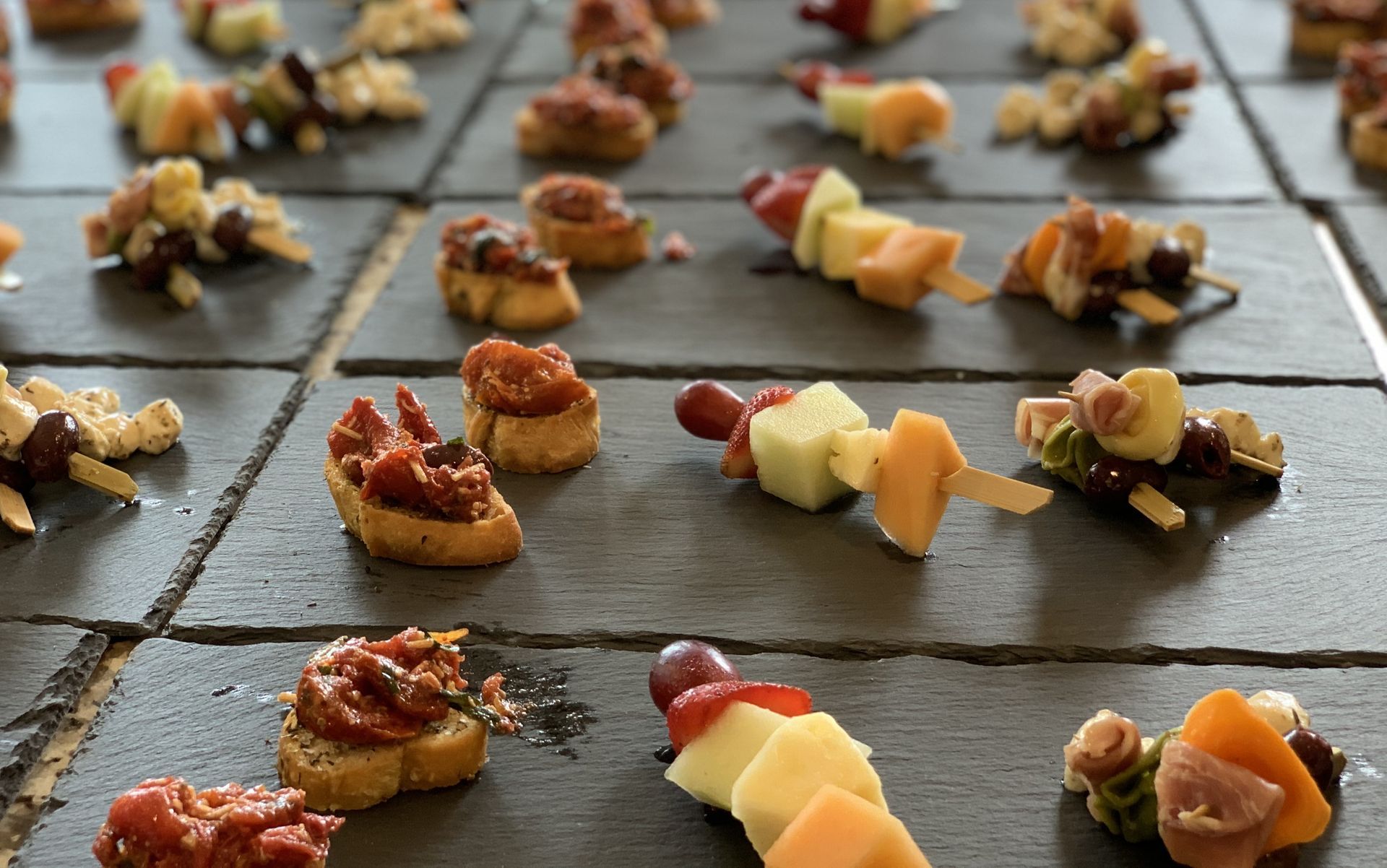 An assortment of bite-sized appetizers, including fruit skewers and bruschetta, arranged on dark slate serving tiles.
