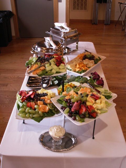 A buffet table covered with a white cloth features several platters of fresh fruit, cheese, and a bowl of dip.