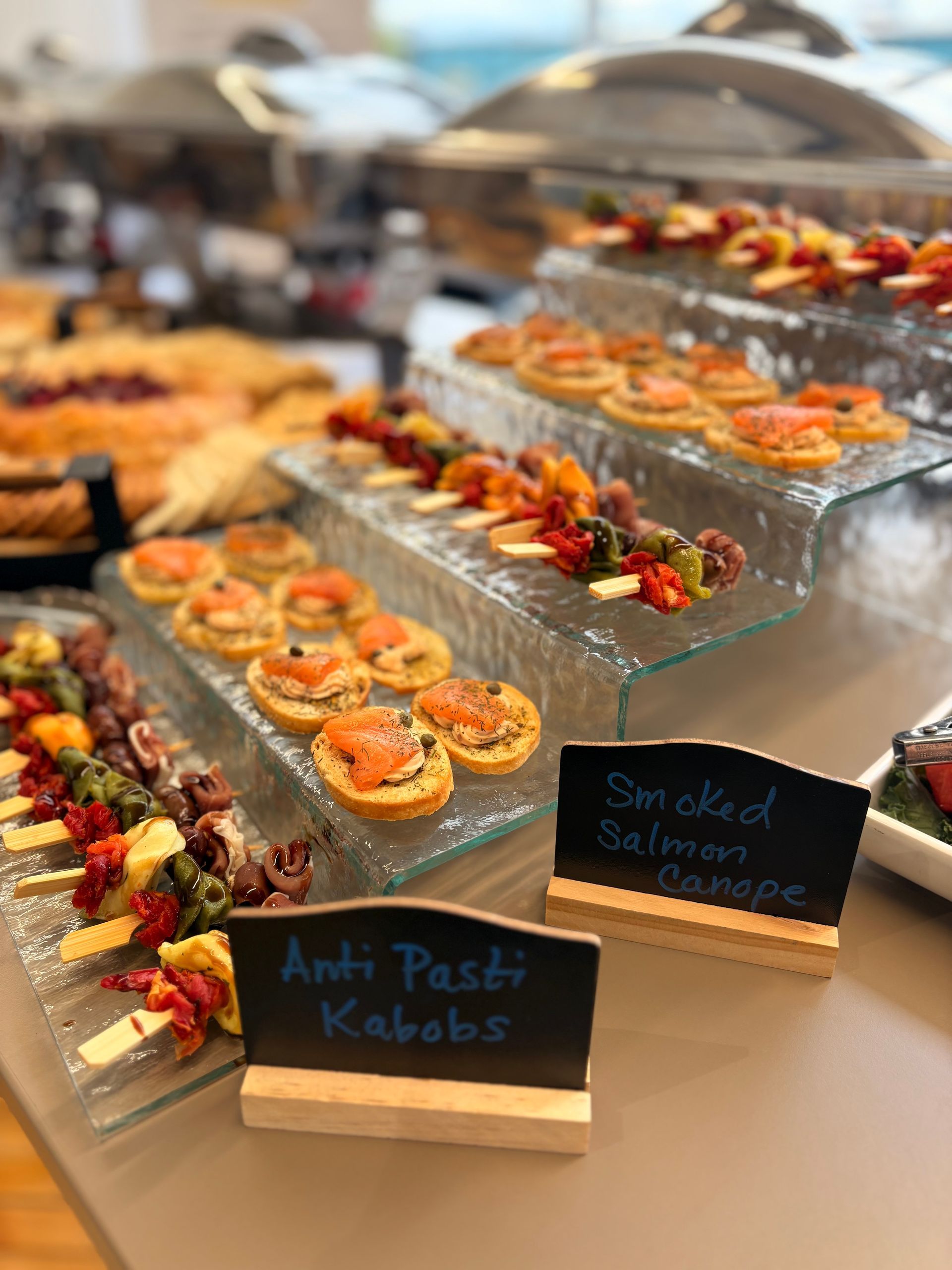 A buffet spread with antipasti kabobs and smoked salmon canapés arranged on glass tiered displays with labeled signs.