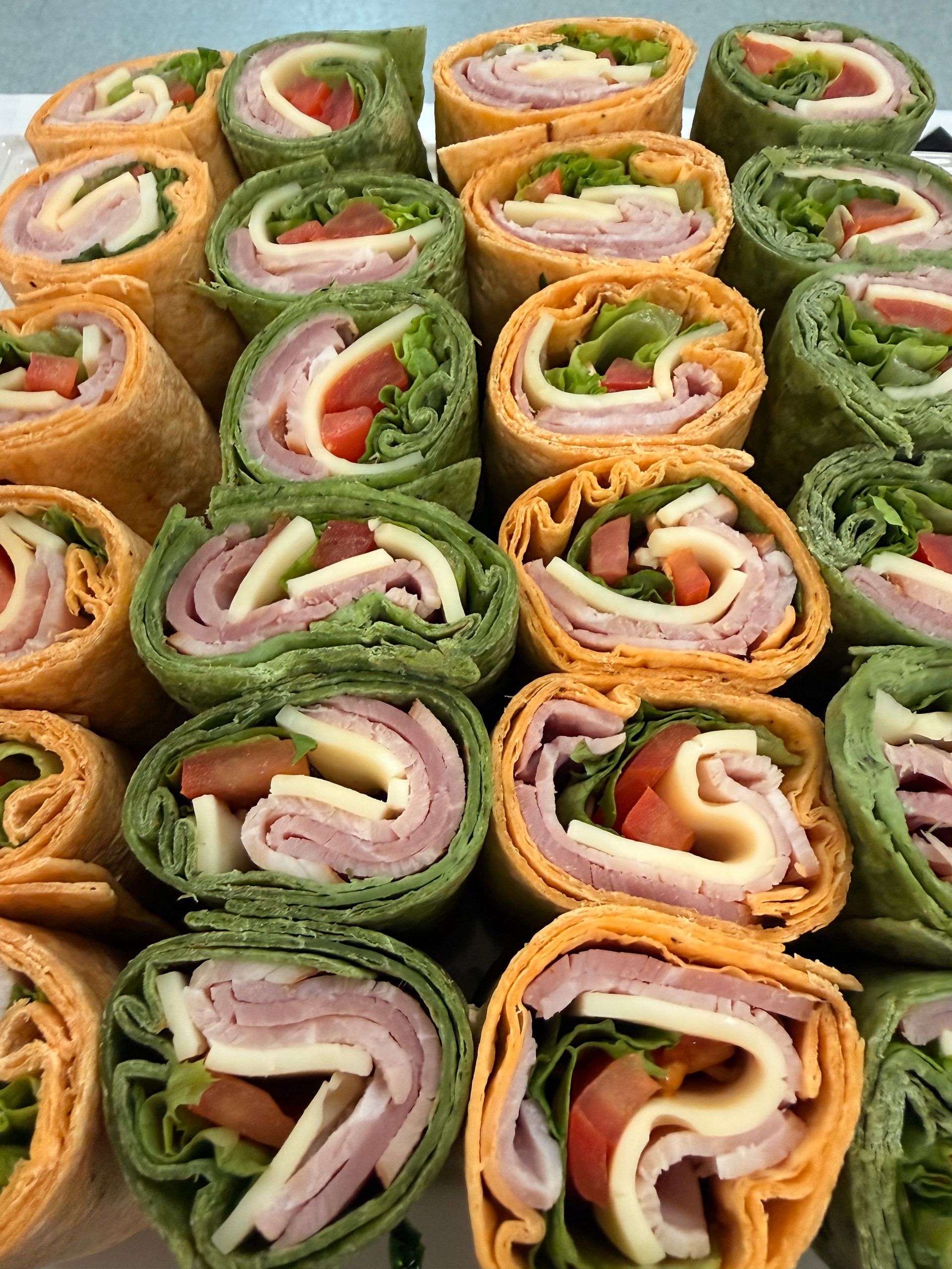 A close-up of a platter of rolled tortilla wraps with ham, cheese, and lettuce, featuring both sun-dried tomato and spinach.