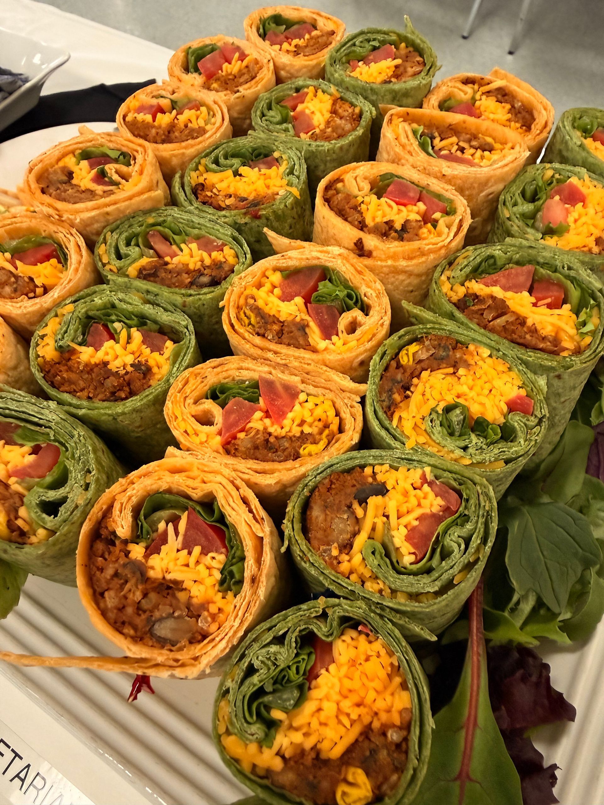 A platter of pinwheel wraps filled with meat, shredded cheddar cheese, and diced tomatoes on a bed of fresh greens.