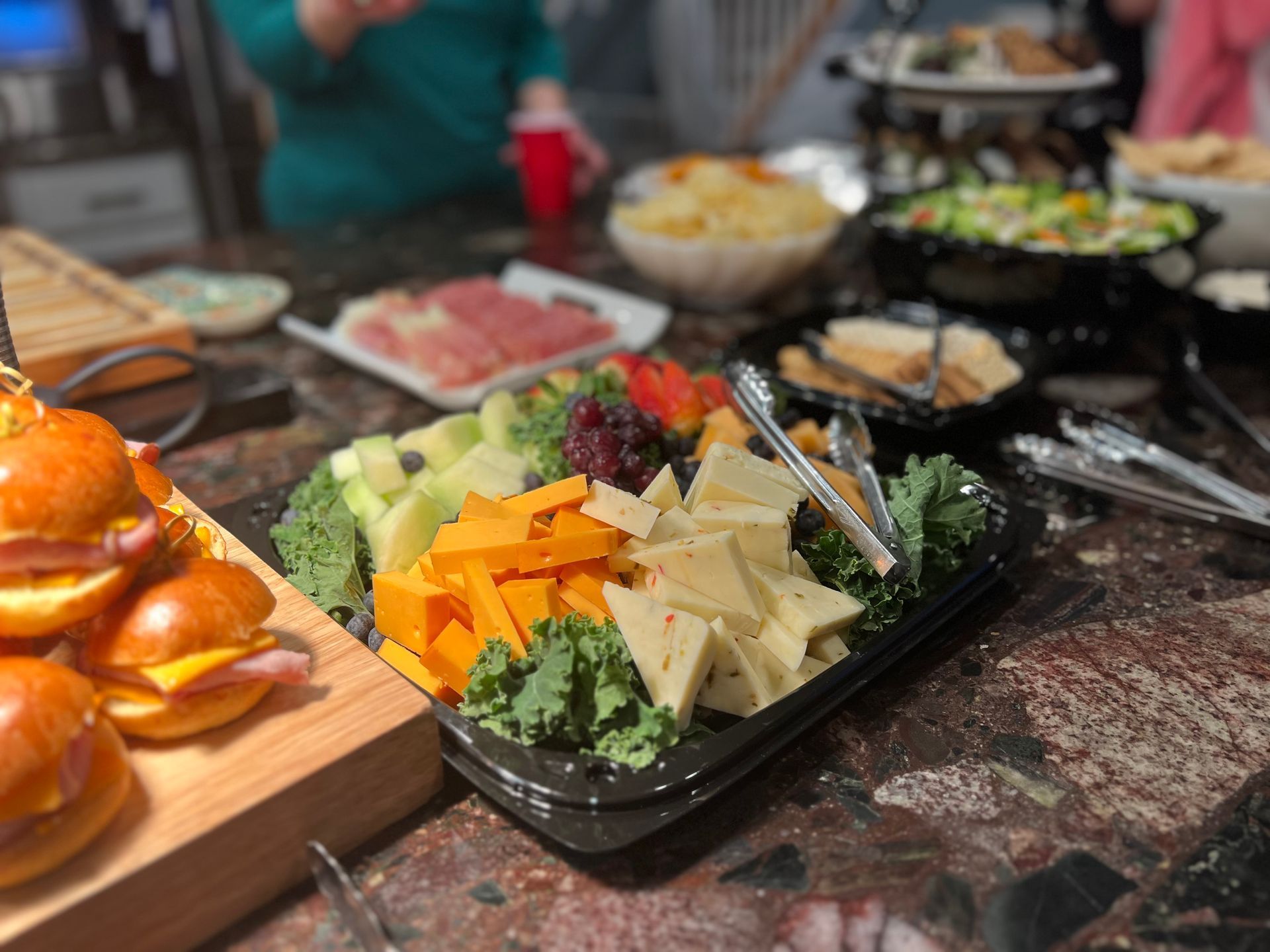 A spread of appetizers on a marble countertop, featuring sliders on a wooden board and a tray of cheese and melon.