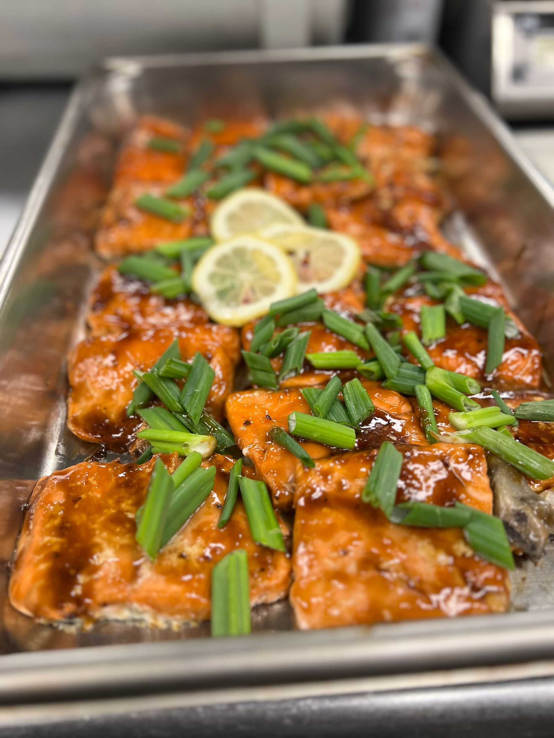 A metal tray filled with glazed salmon fillets, garnished with chopped green onions and two lemon slices.