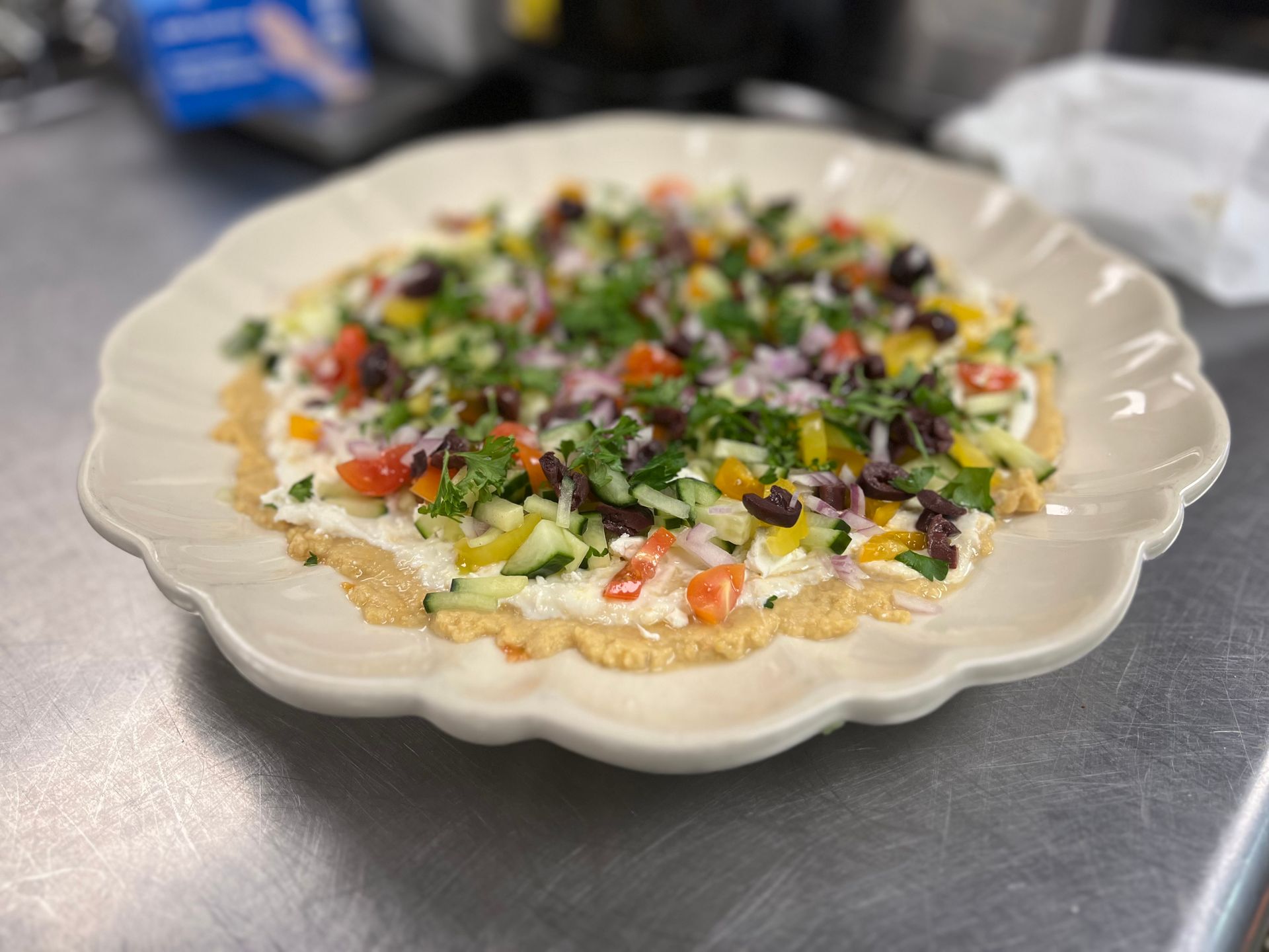 A colorful Mediterranean veggie pizza topped with chopped cucumbers, tomatoes, peppers, onions, and olives on a platter.