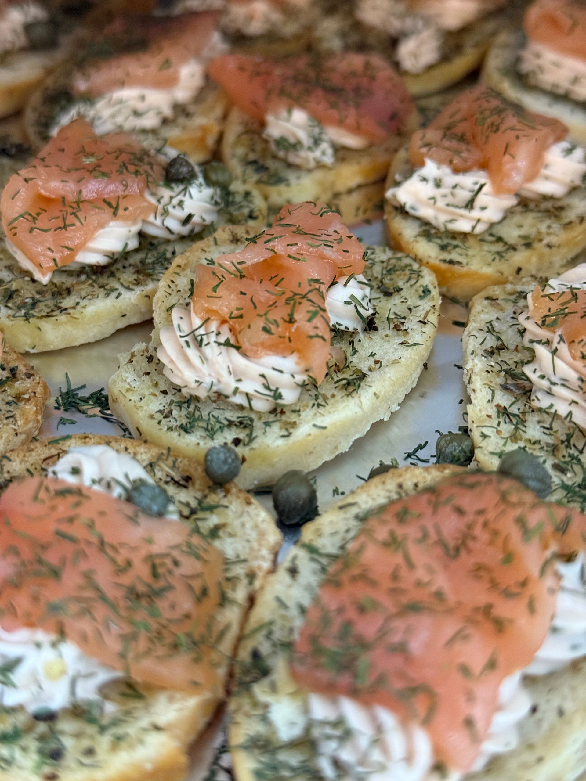 Close-up of several toasted bread slices topped with cream cheese, smoked salmon, dill, and capers on a tray.