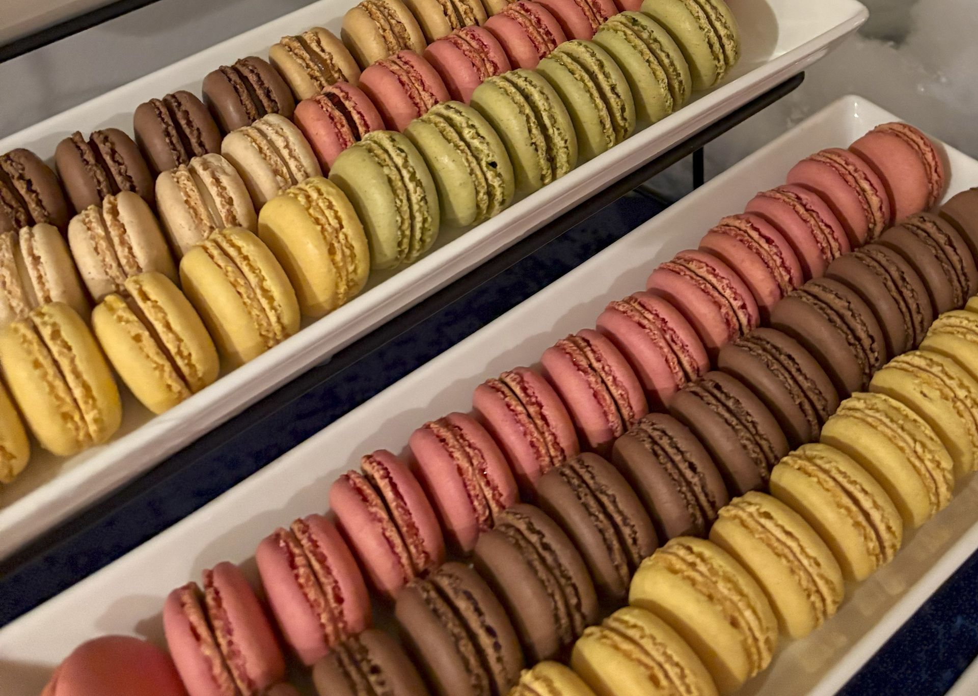Two white rectangular serving platters filled with rows of assorted colorful macarons, including pink, brown, and yellow.