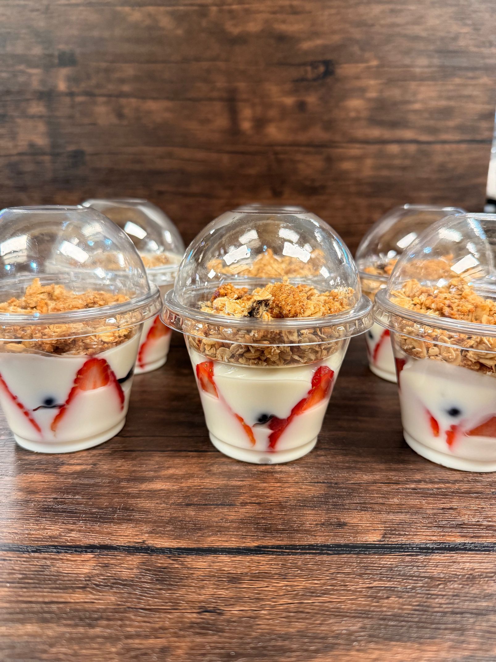Several clear plastic cups filled with yogurt, sliced strawberries, and granola, arranged on a dark wooden surface.
