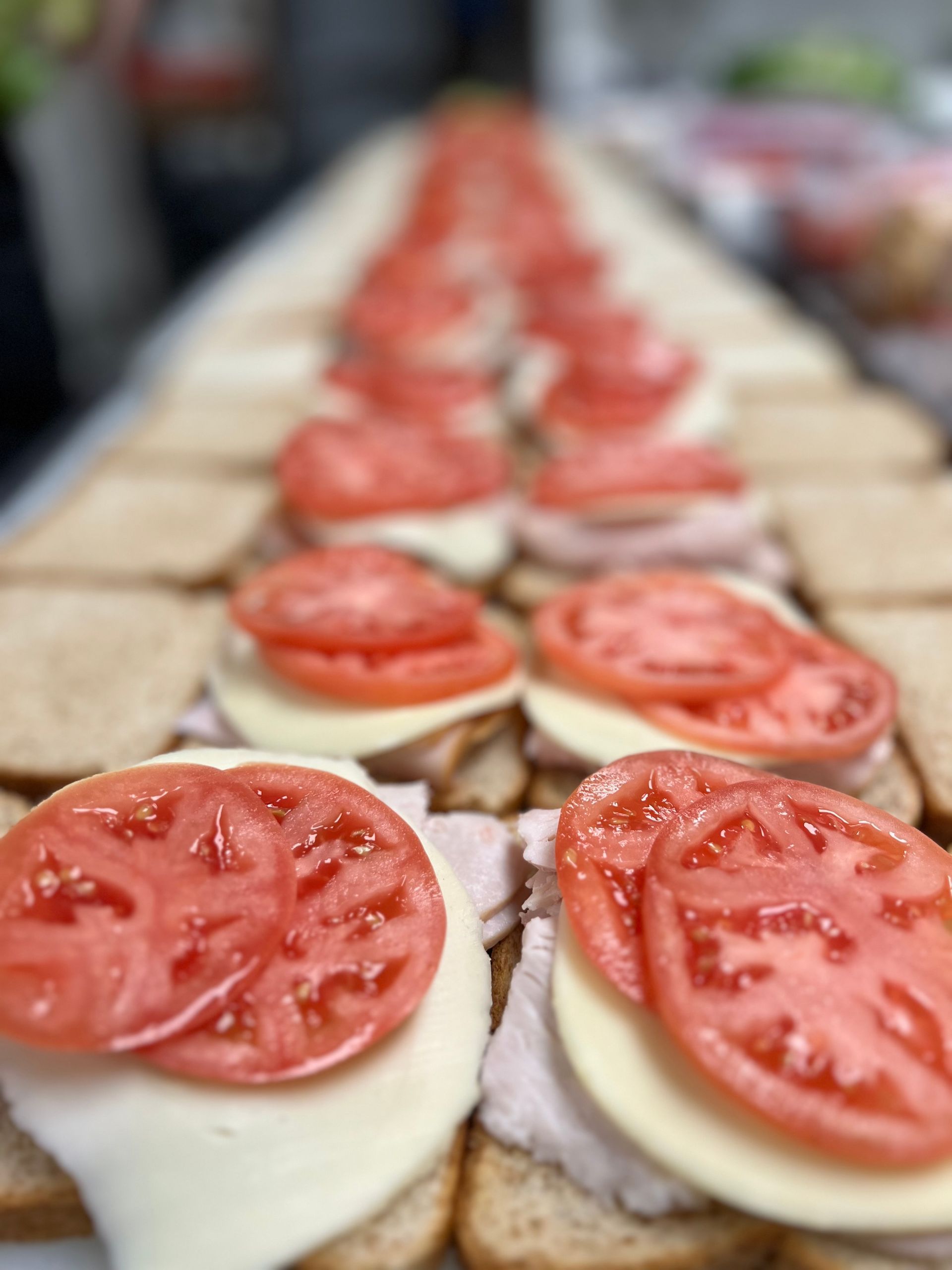 Multiple slices of bread lined up on a counter, each topped with a slice of cheese, deli meat, and fresh tomato slices.