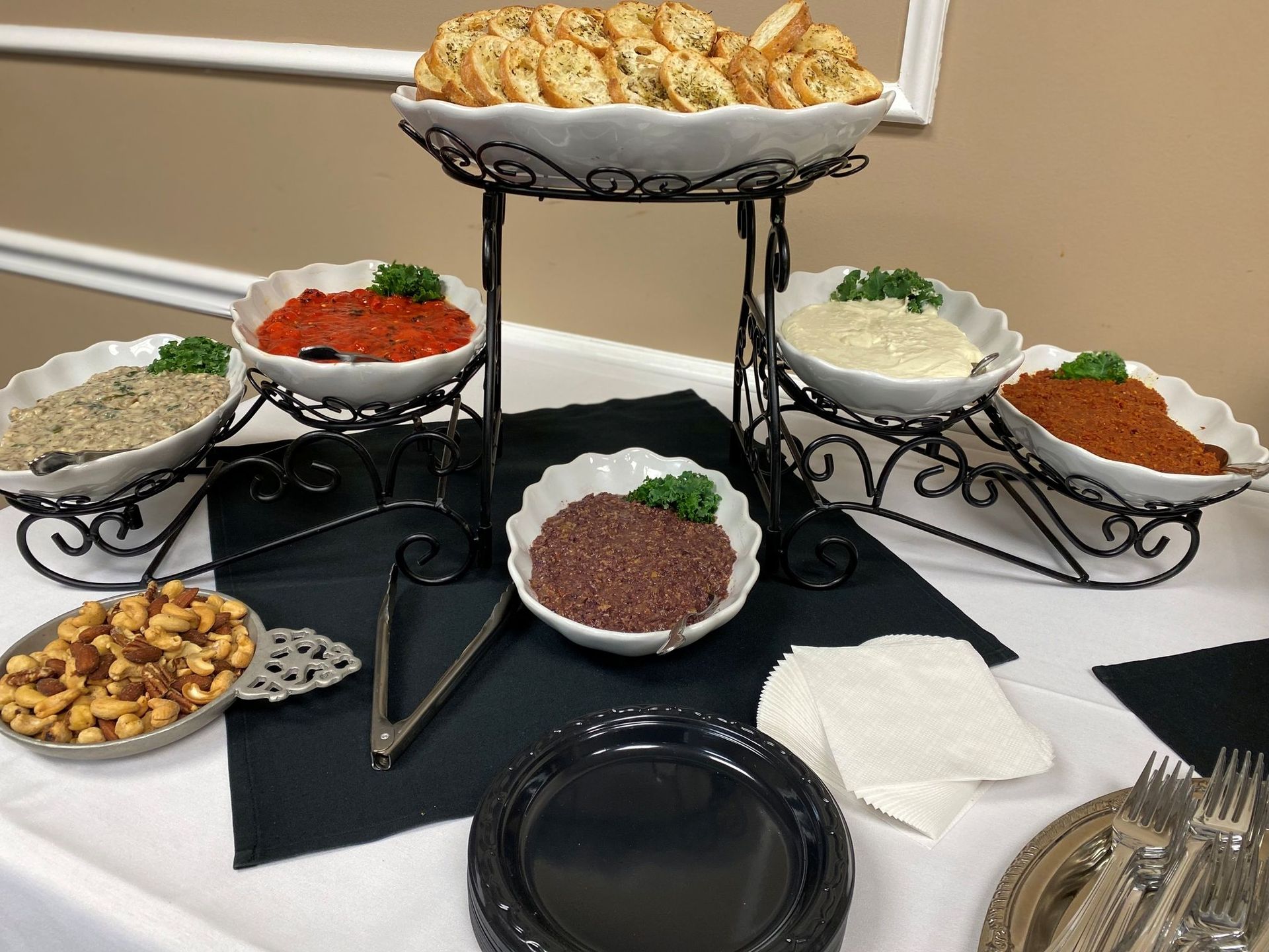 Appetizer spread with dips, crackers, and olives on a tiered stand and a table set with plates and cutlery.