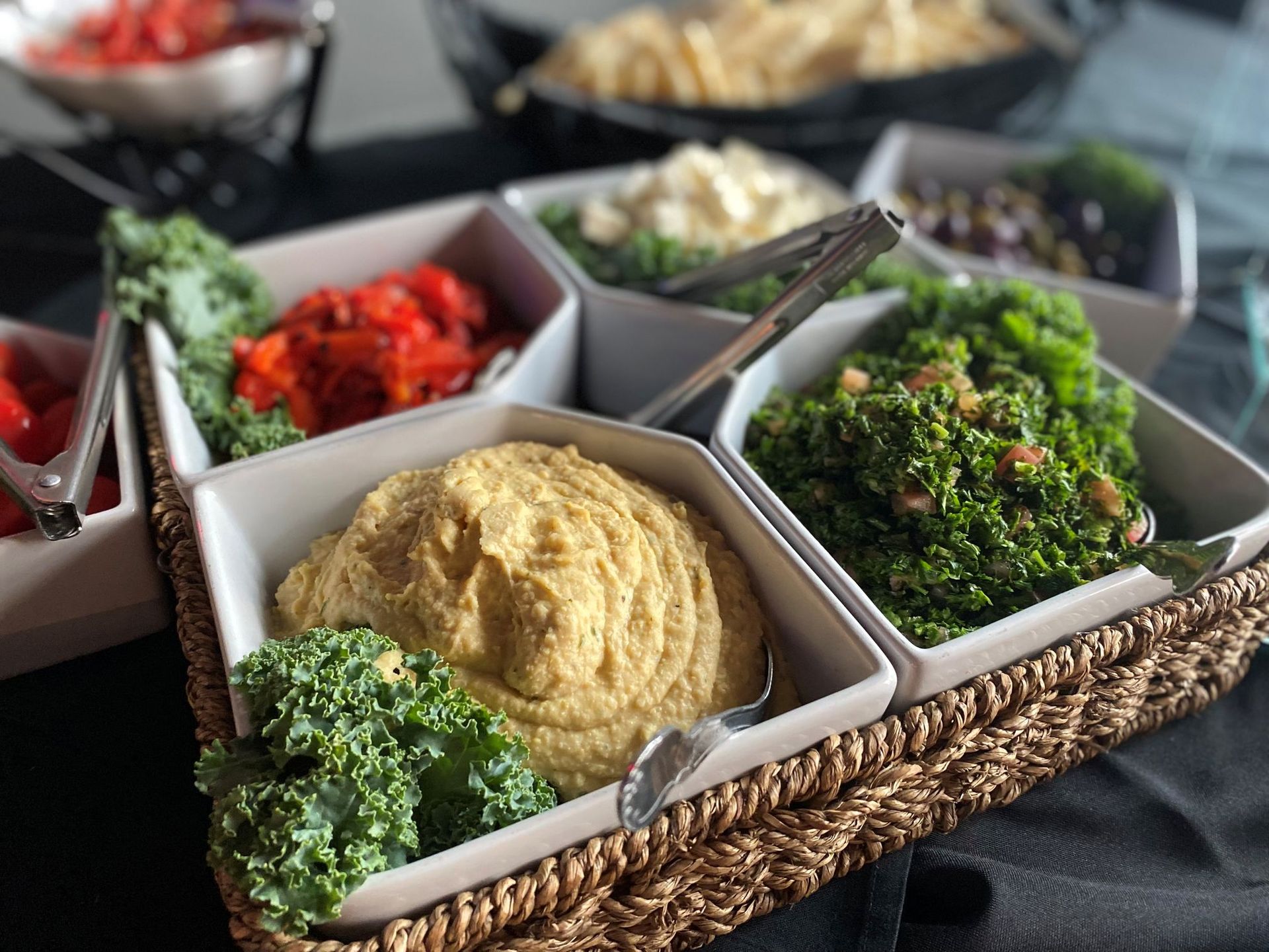 A woven basket filled with white dishes containing hummus, roasted vegetables, and olives.