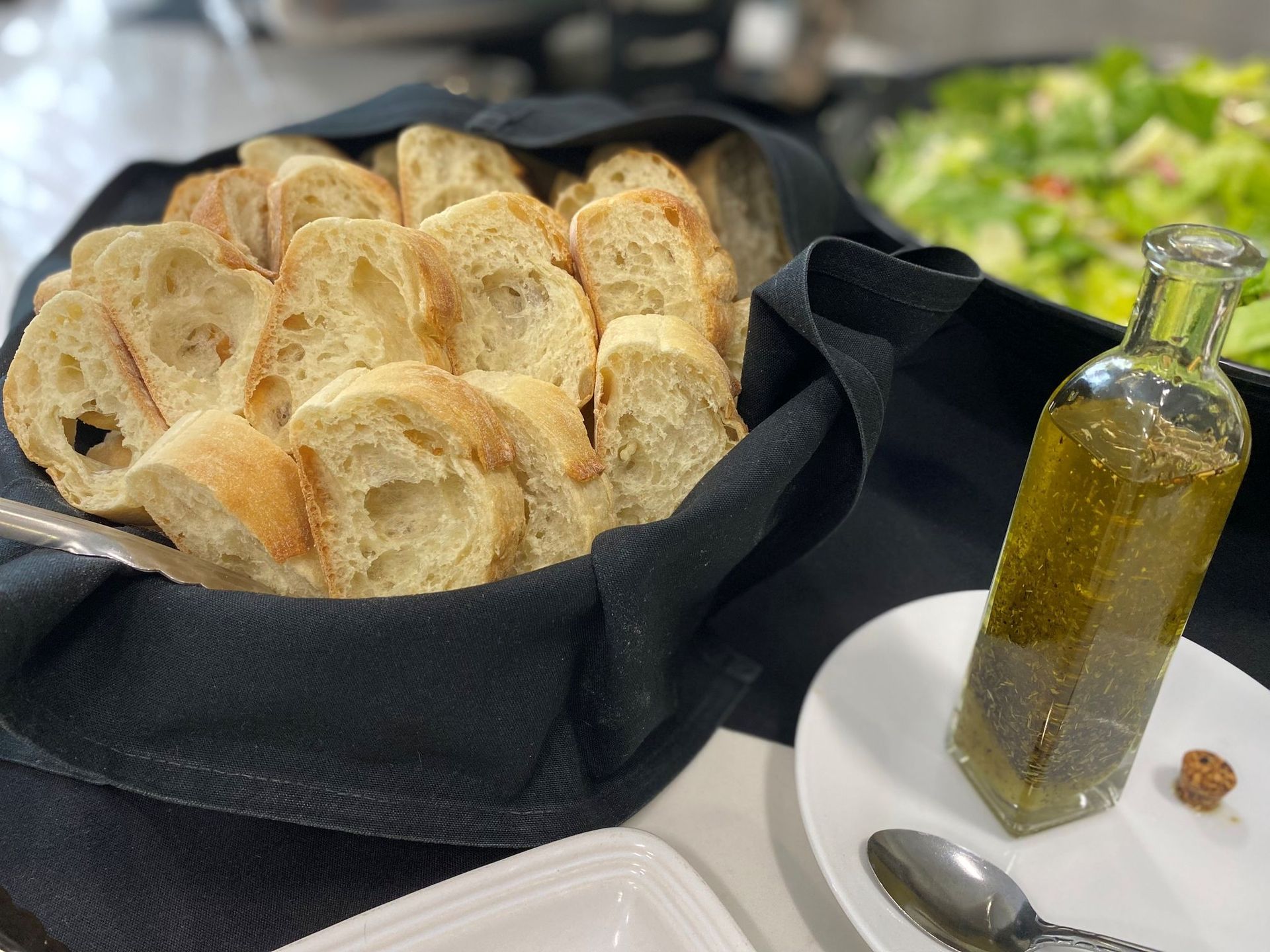 Basket of bread slices, bottle of olive oil with herbs, and salad in a buffet setting.