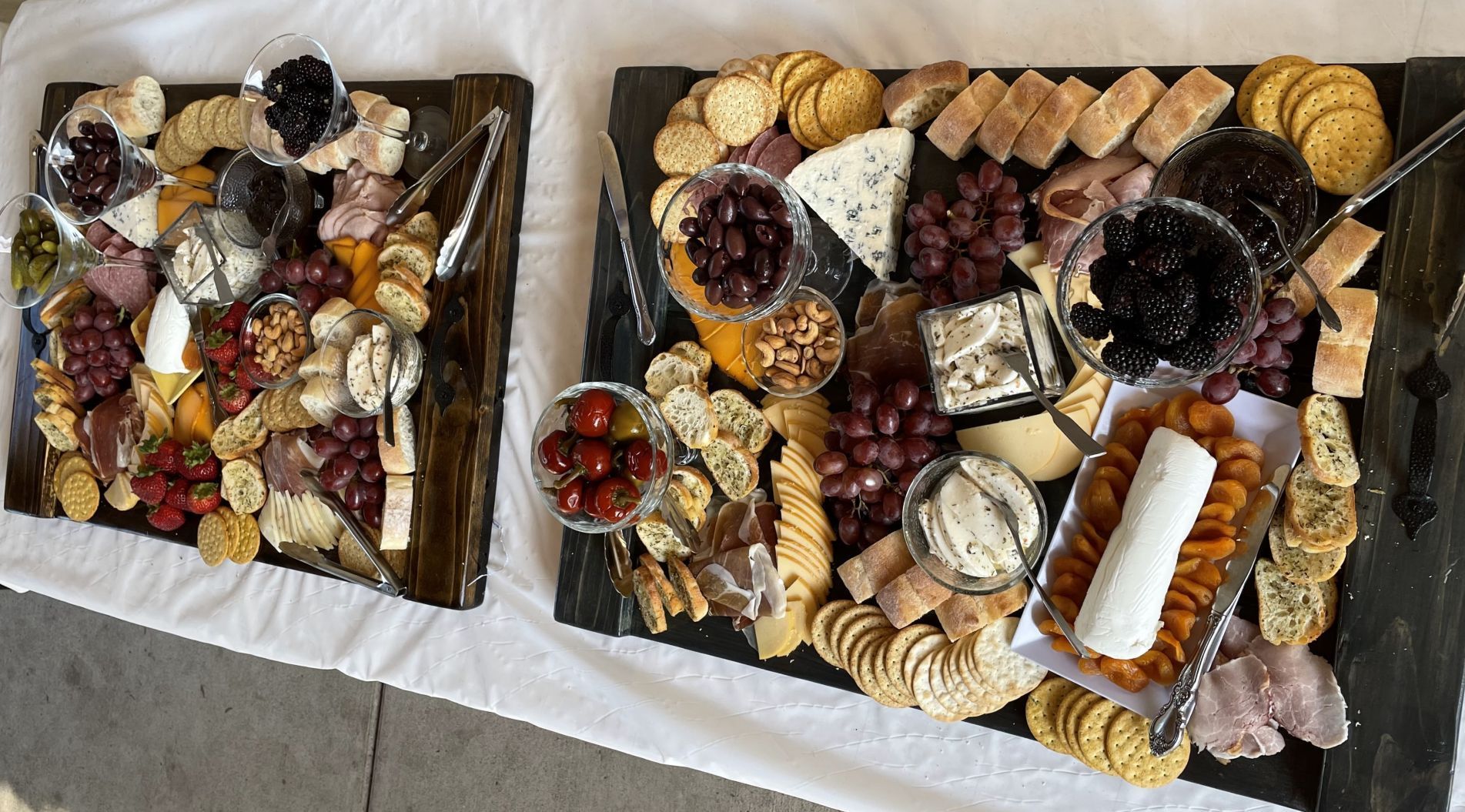 Two charcuterie boards with various snacks, including cheeses, crackers, fruits, and meats, arranged on a white tablecloth.