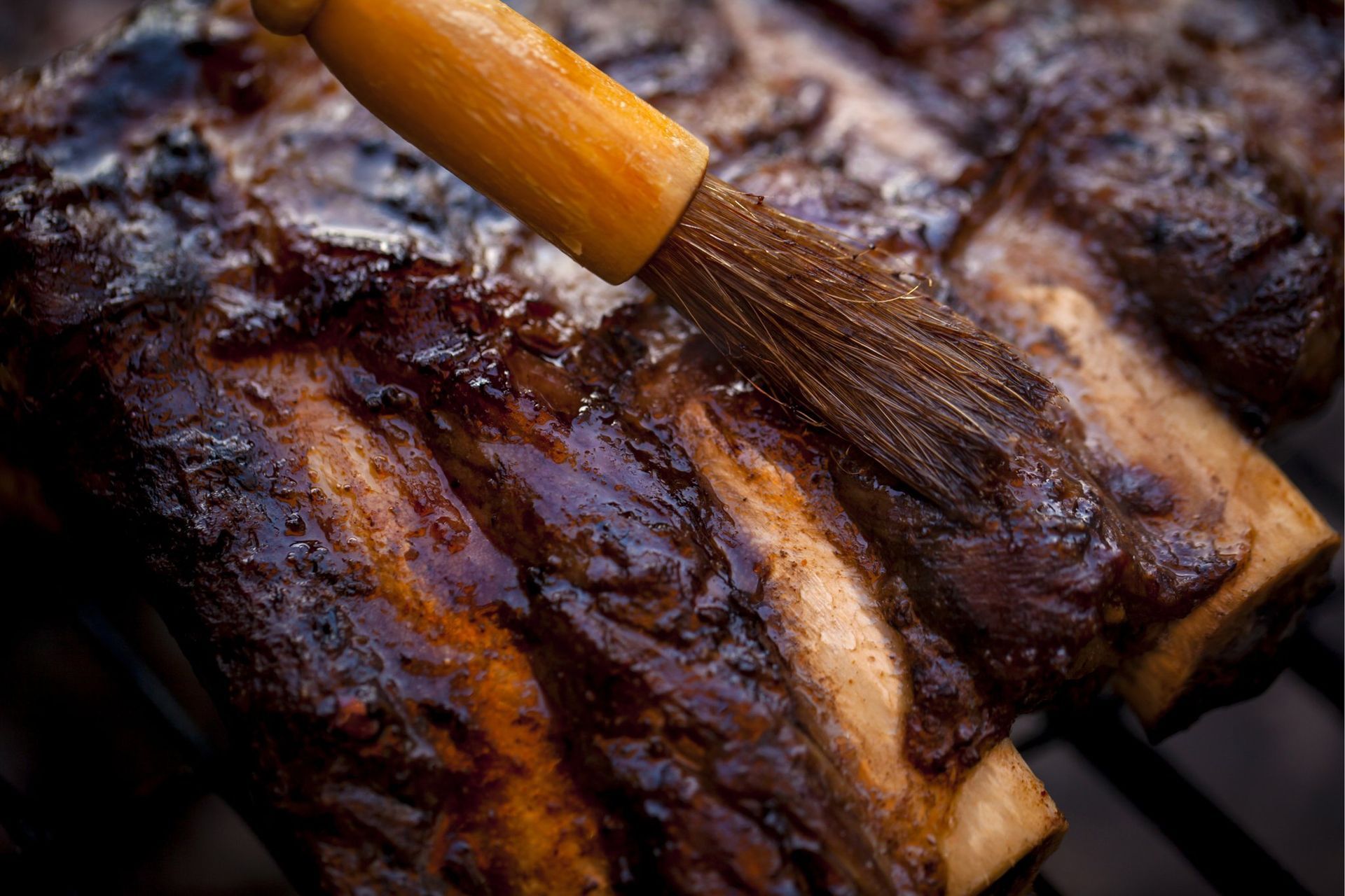 Ribs on grill, brushed with dark sauce; close-up shows glossy meat, wooden brush.