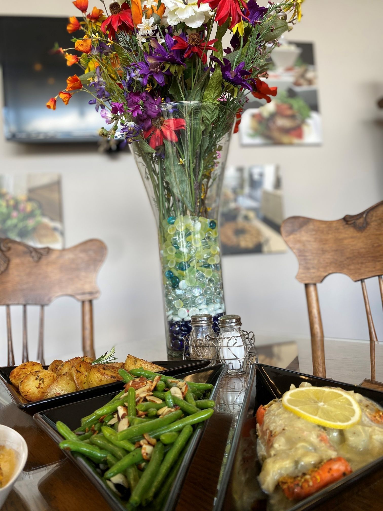 Table set with plates of food, green beans, and a dish with sauce. Vase of flowers in the center.