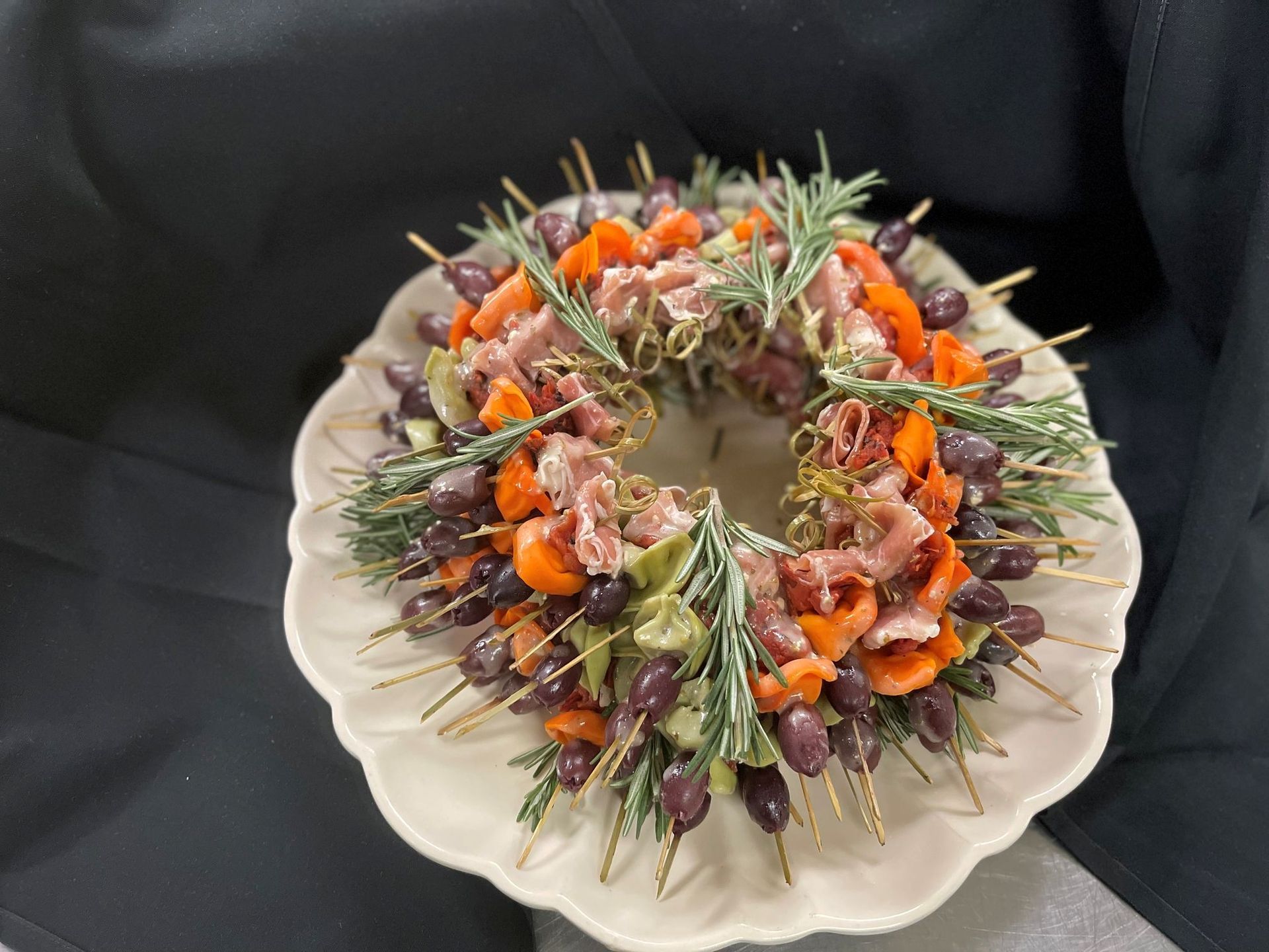 Appetizer wreath on a white plate: olives, prosciutto, apricots, and rosemary on skewers.
