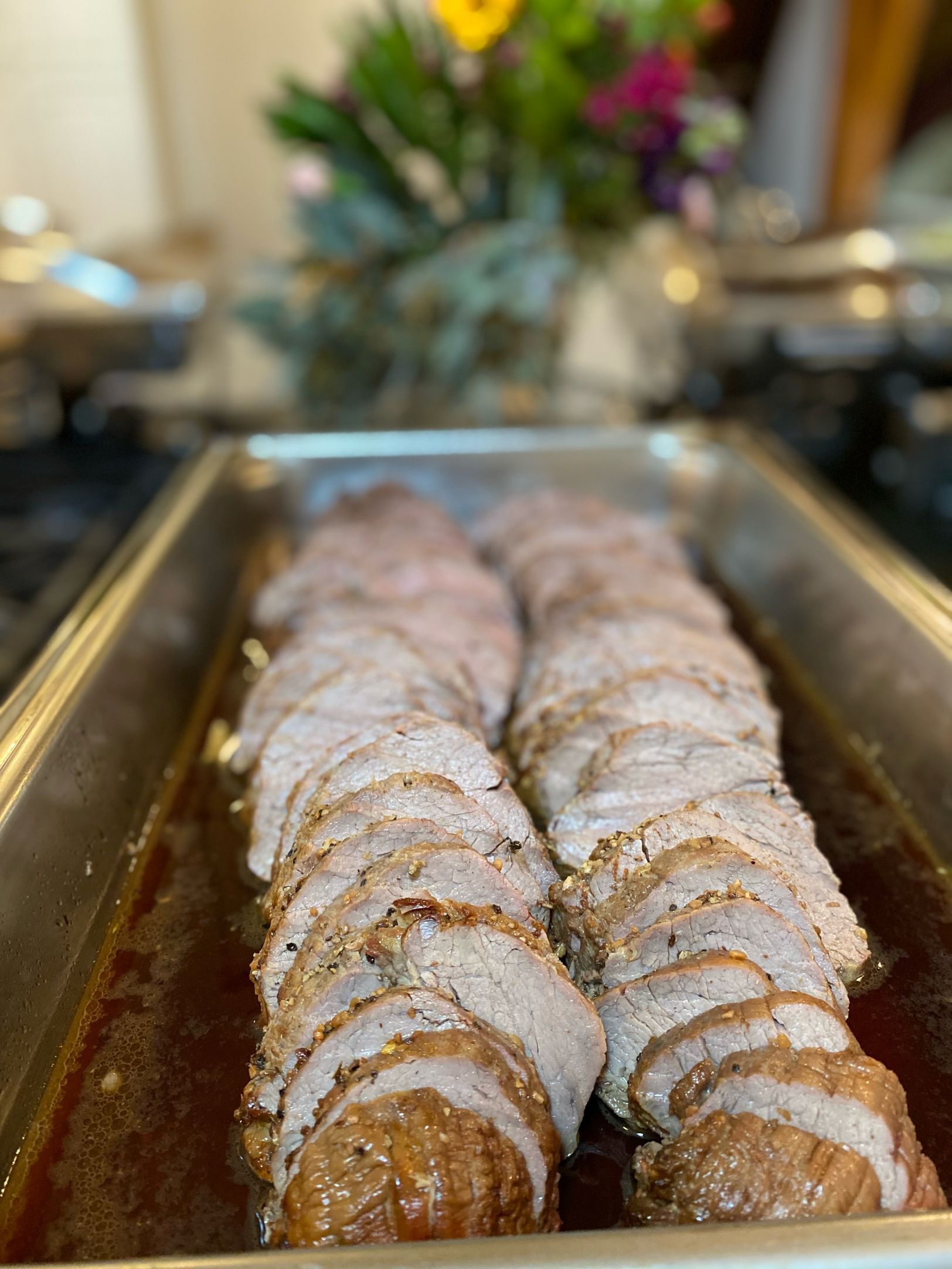 Sliced roast pork in a metal pan, seasoned, with a dark sauce. Blurred floral arrangement in background.