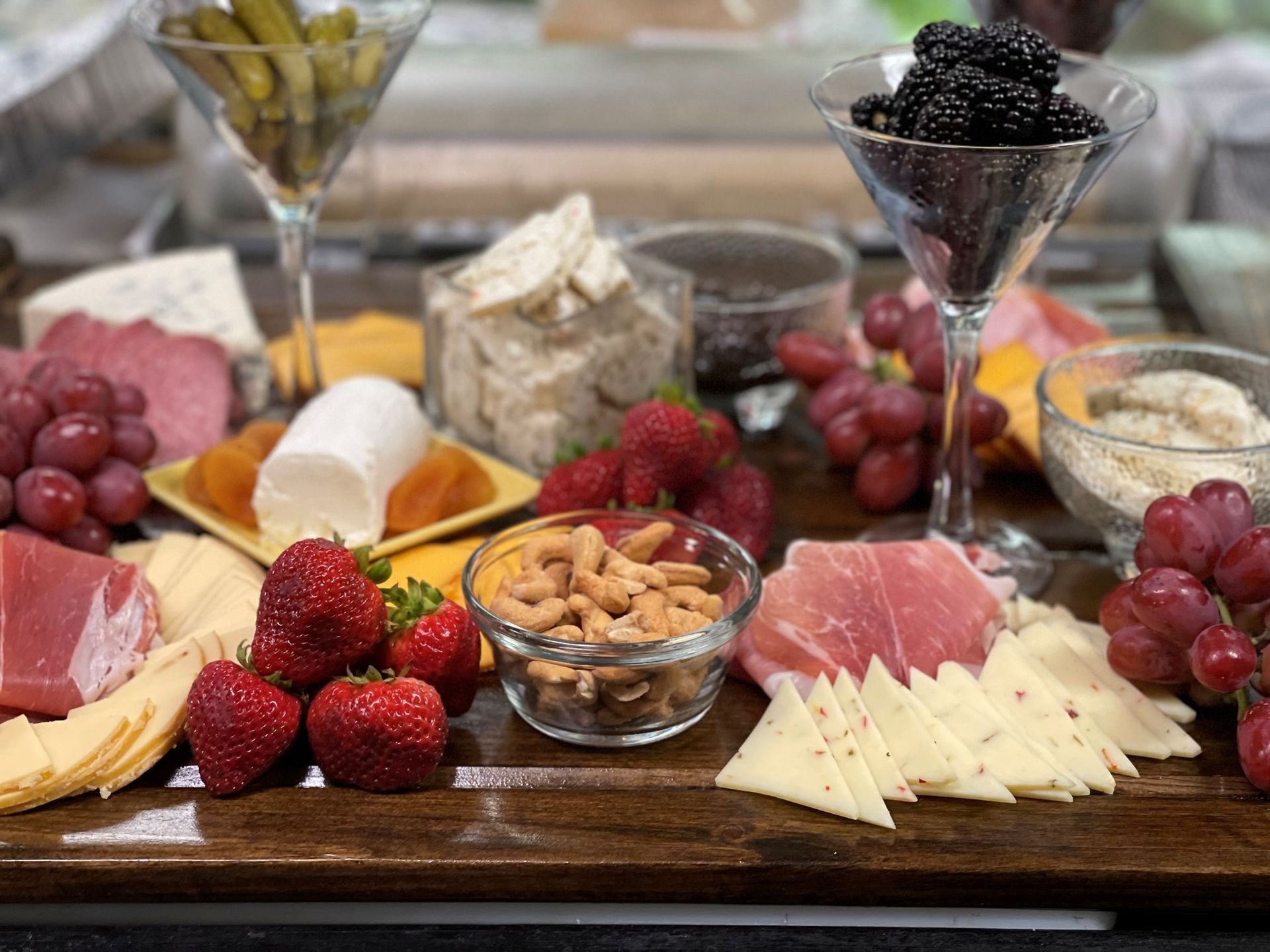 Charcuterie board with various cheeses, fruits, nuts, crackers, pickles, and berries, in cocktail glasses.