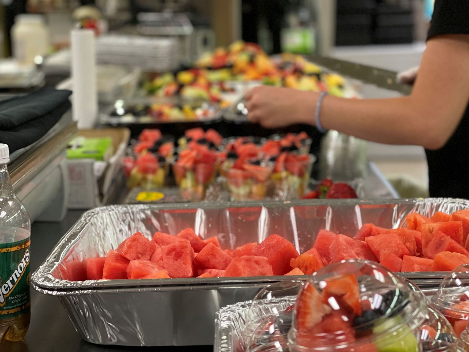 Fruit buffet: Cubed watermelon in trays, hands preparing fruit cups, various colorful fruits.