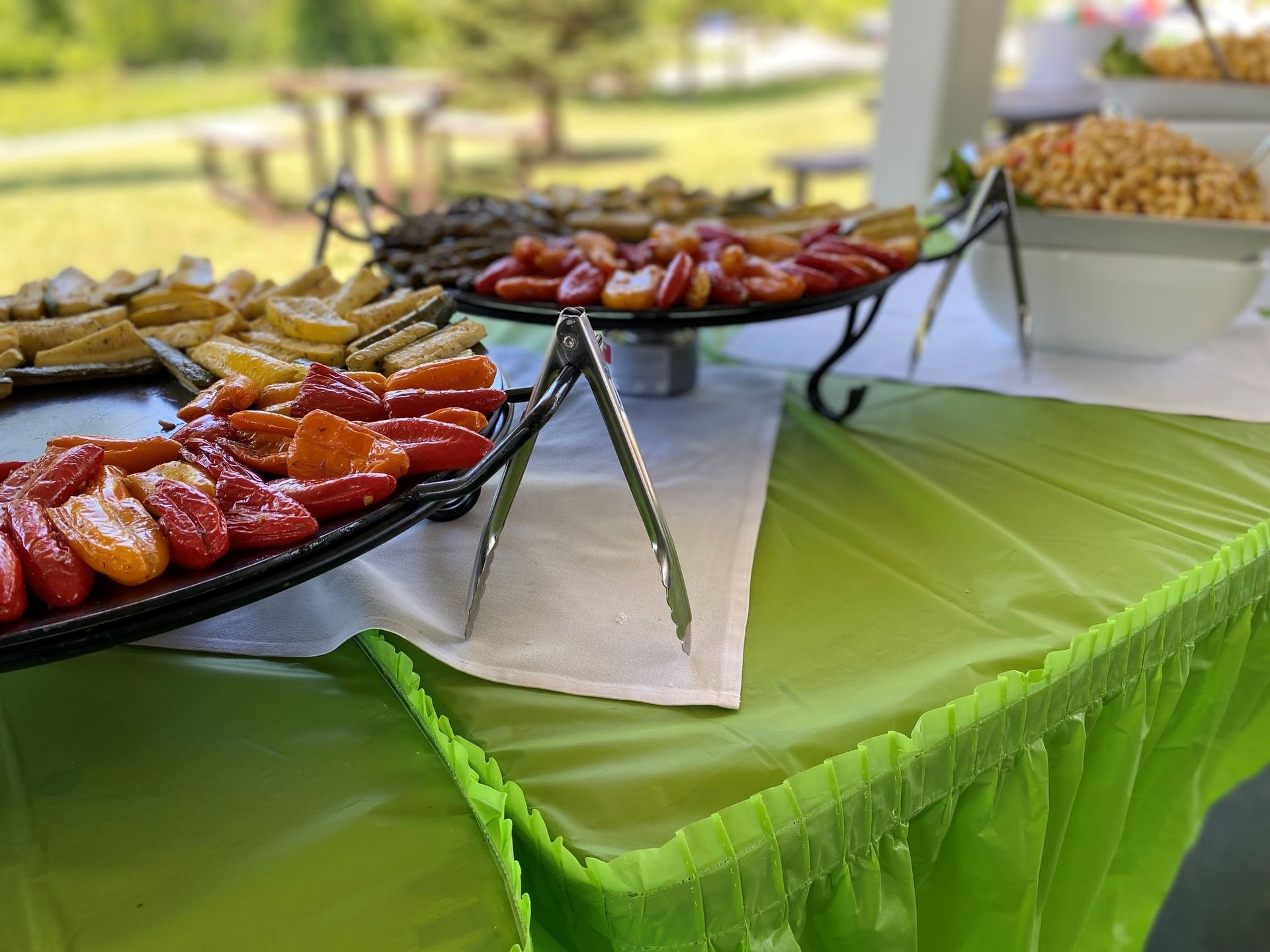 Buffet table with colorful peppers and other dishes, outdoors with picnic tables in the background.