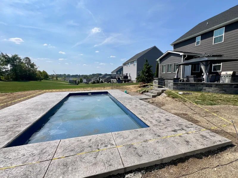 Rectangular pool under construction with concrete patio, near a house and grassy field, on a sunny day.