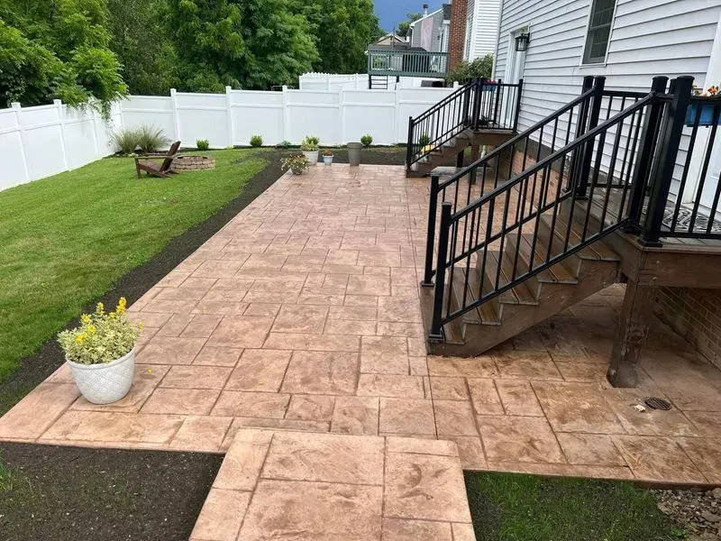 Stamped concrete patio with steps and black railing, surrounded by grass and a white fence.