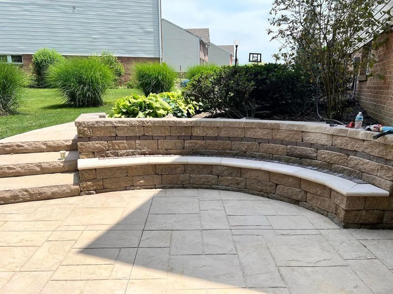 A curved stone bench on a patio. Steps on the left lead to grass and shrubs.