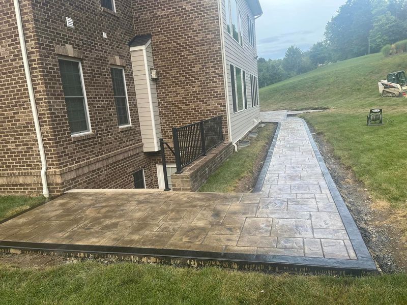 Brick house with concrete patio and pathway, bordered by lawn, leading up hill.