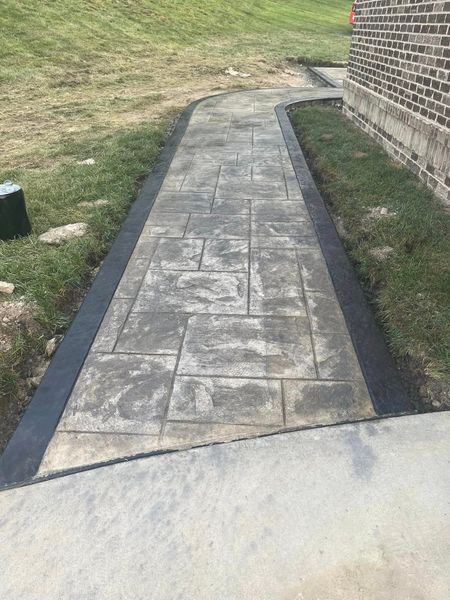 Concrete walkway with textured gray blocks and dark border, next to a brick building and grass.
