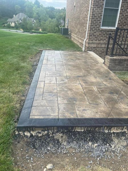Concrete patio with stone pattern and dark border next to a brick building and grass.