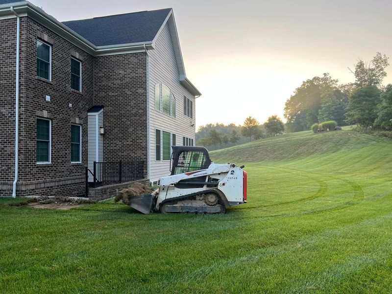 A Bobcat compact track loader on a lawn next to a two-story brick house, under a sunrise.