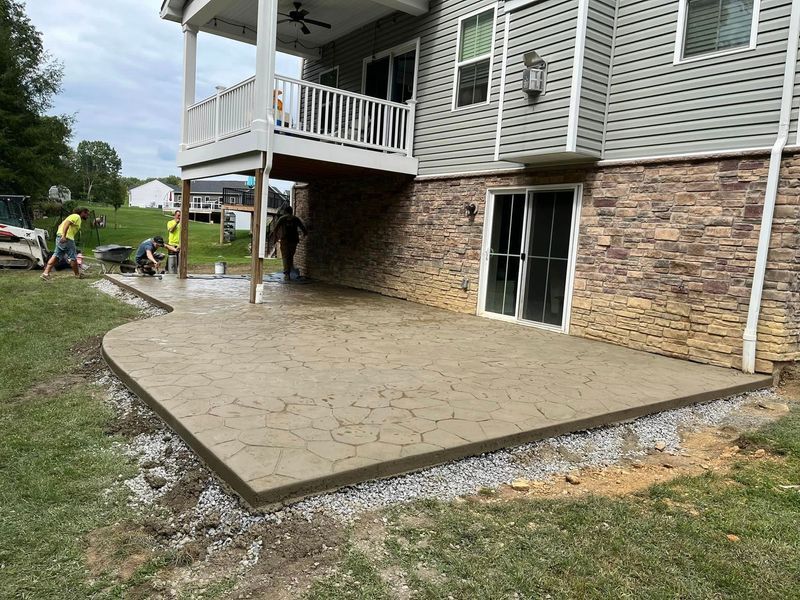 Workers pouring concrete patio under a house's elevated deck. Gray concrete with decorative texture, grassy yard, and gravel edging.