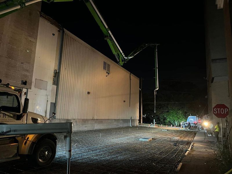 Concrete pour at night; concrete pump arm extends over a building. Men work near a stop sign.