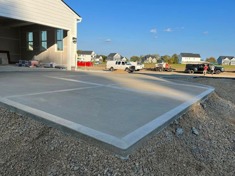 Newly poured concrete driveway next to a building under construction, with gravel and vehicles in the background.