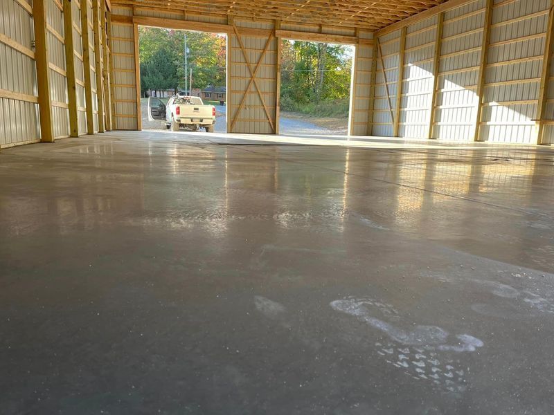 Interior of a large open building with a newly poured, wet concrete floor. A truck is visible through open doors.